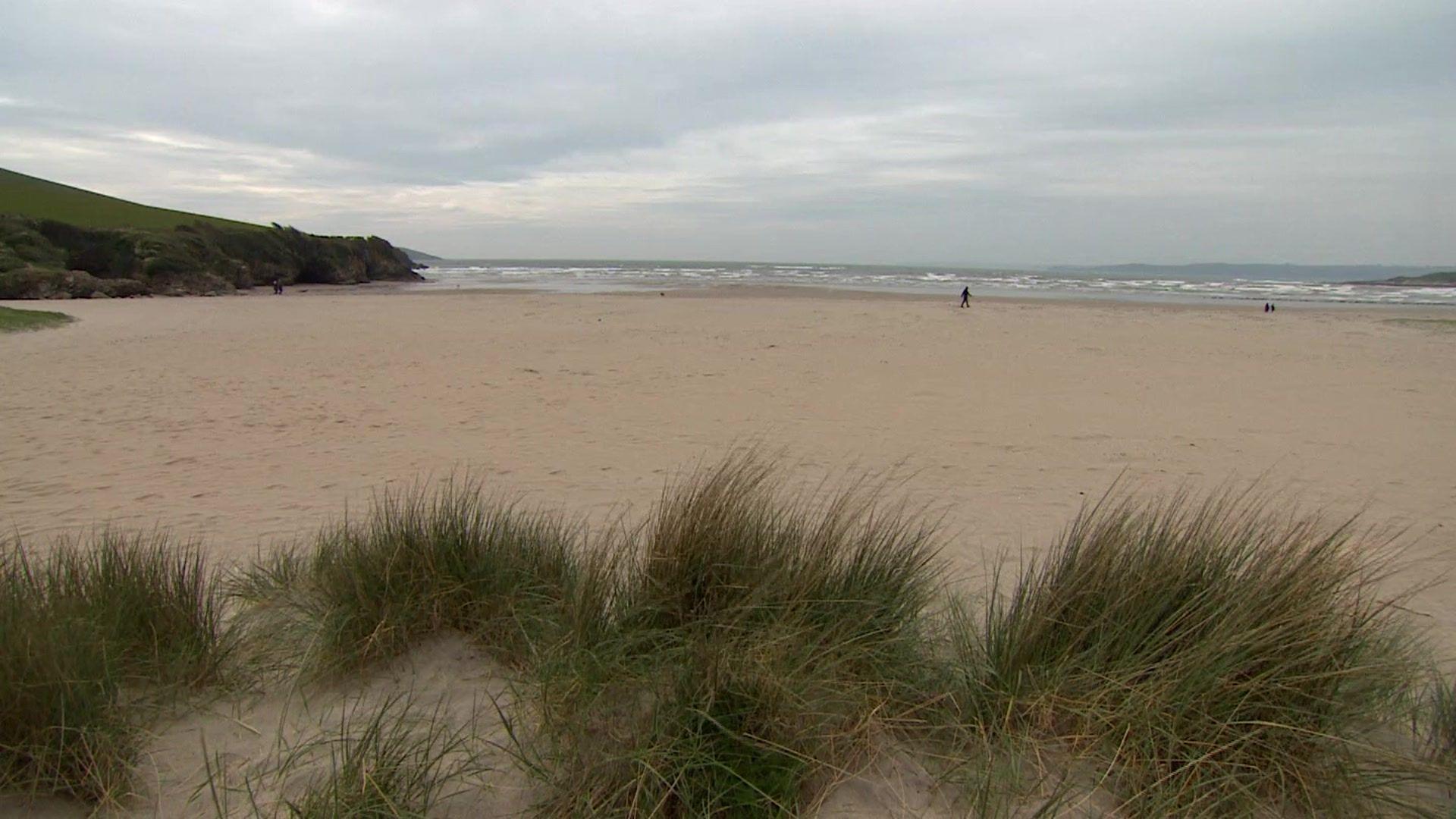 A wide expanse of sand on a blustery day with sand dunes and grasses visible in the foreground and a loner walker in the background near the ocean which is grey and has white horses