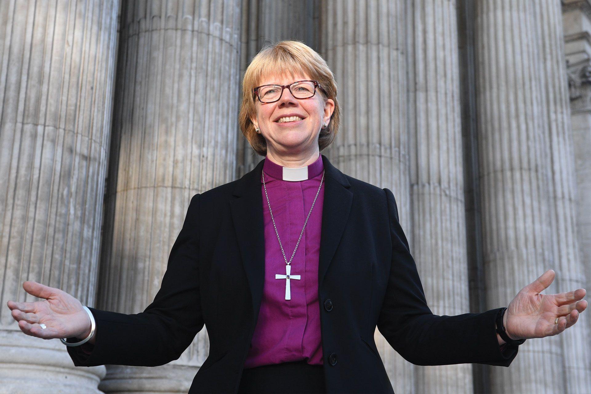 Dame Sarah Mullally is dressed in a dark jacket and is wearing a white clerical collar on a purple shirt. Her arms are outstretched and she is smiling. Large Roman style pillars can be seen in the background. 