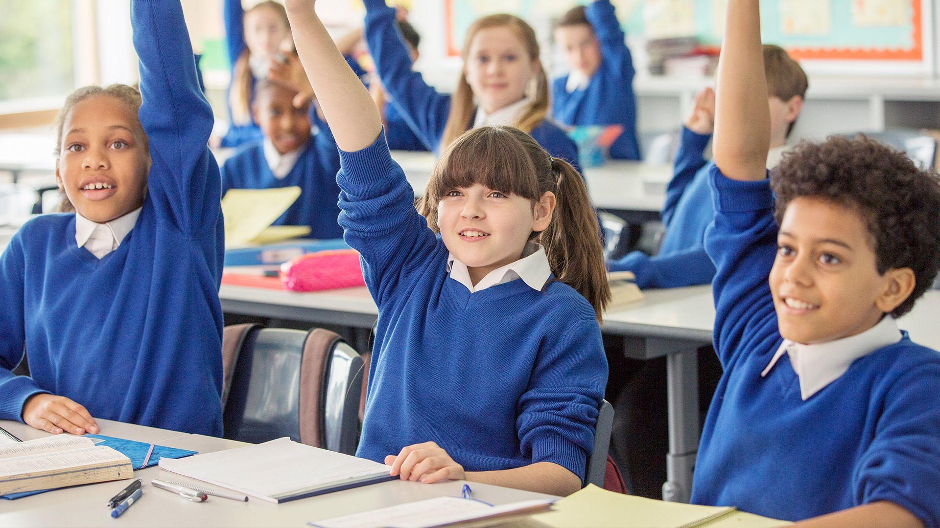 Children sit at their desks in school uniform. They all have their hands up to answer a question