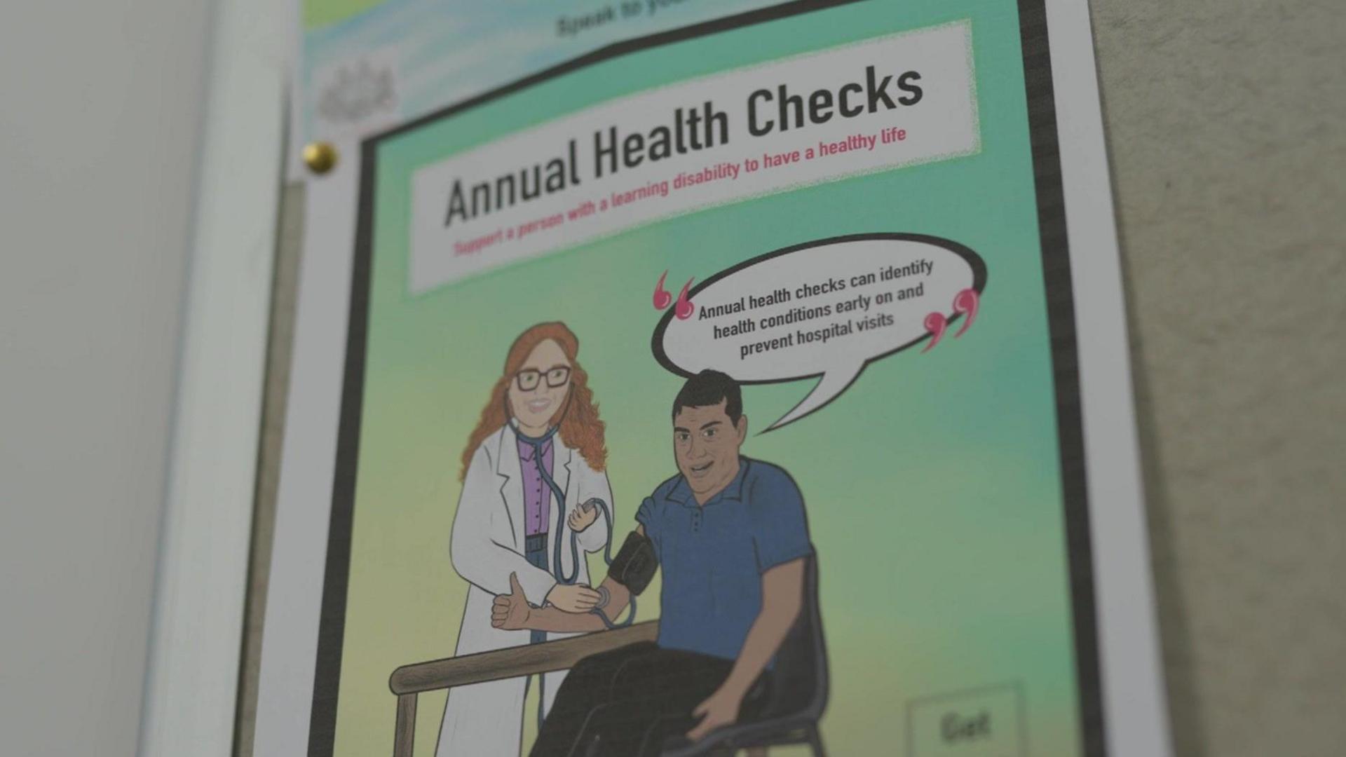 A poster on a wall of a GP practice shows a drawing of a patient having their blood pressure taken by a GP. The text on the picture gives information about annual health checks and how they can prevent hospital visits.