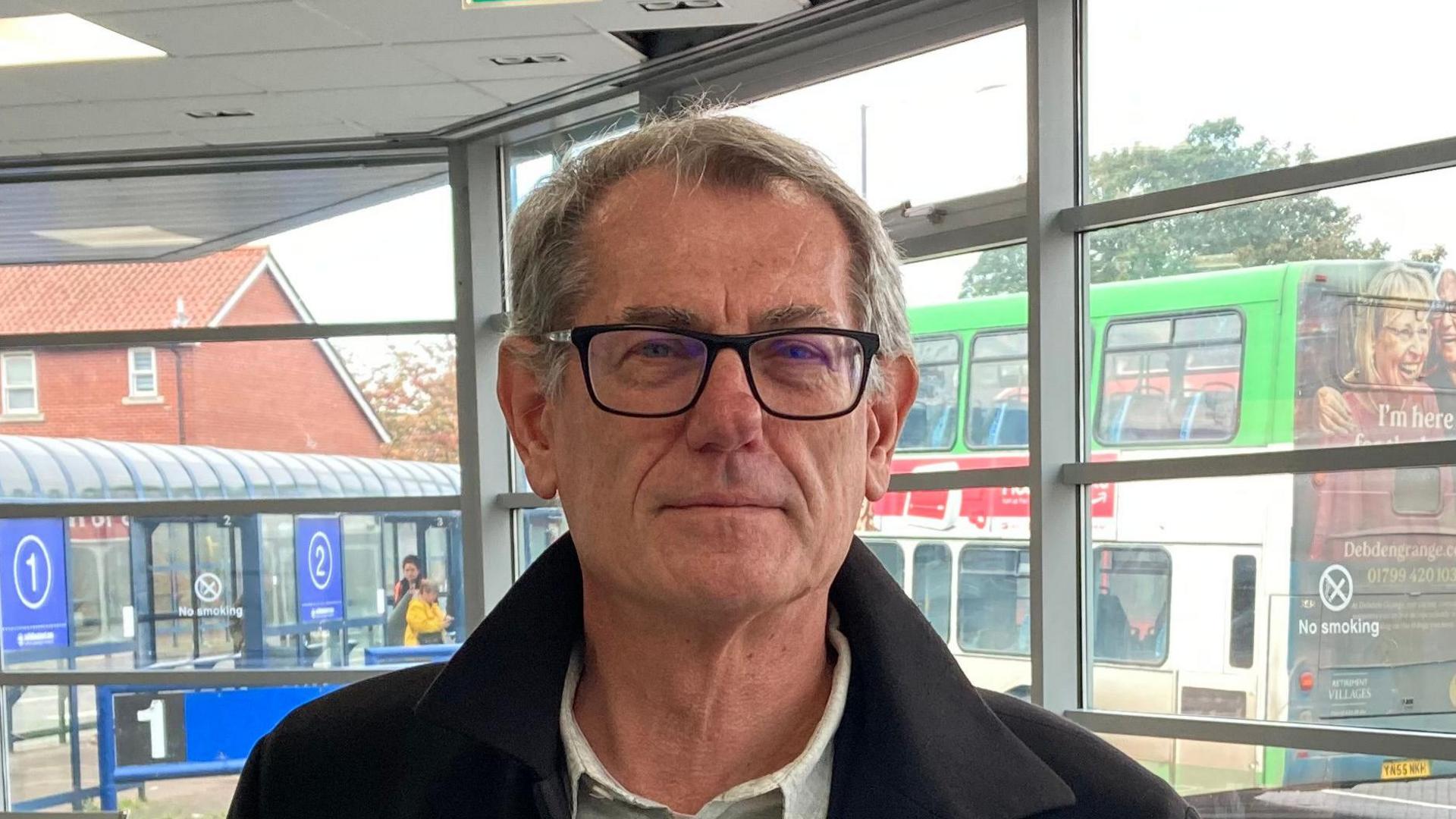 Cliff Waterman smiles at the camera while standing in a bus station. He has short grey hair. He wears black glasses and a black coat with a shirt underneath.