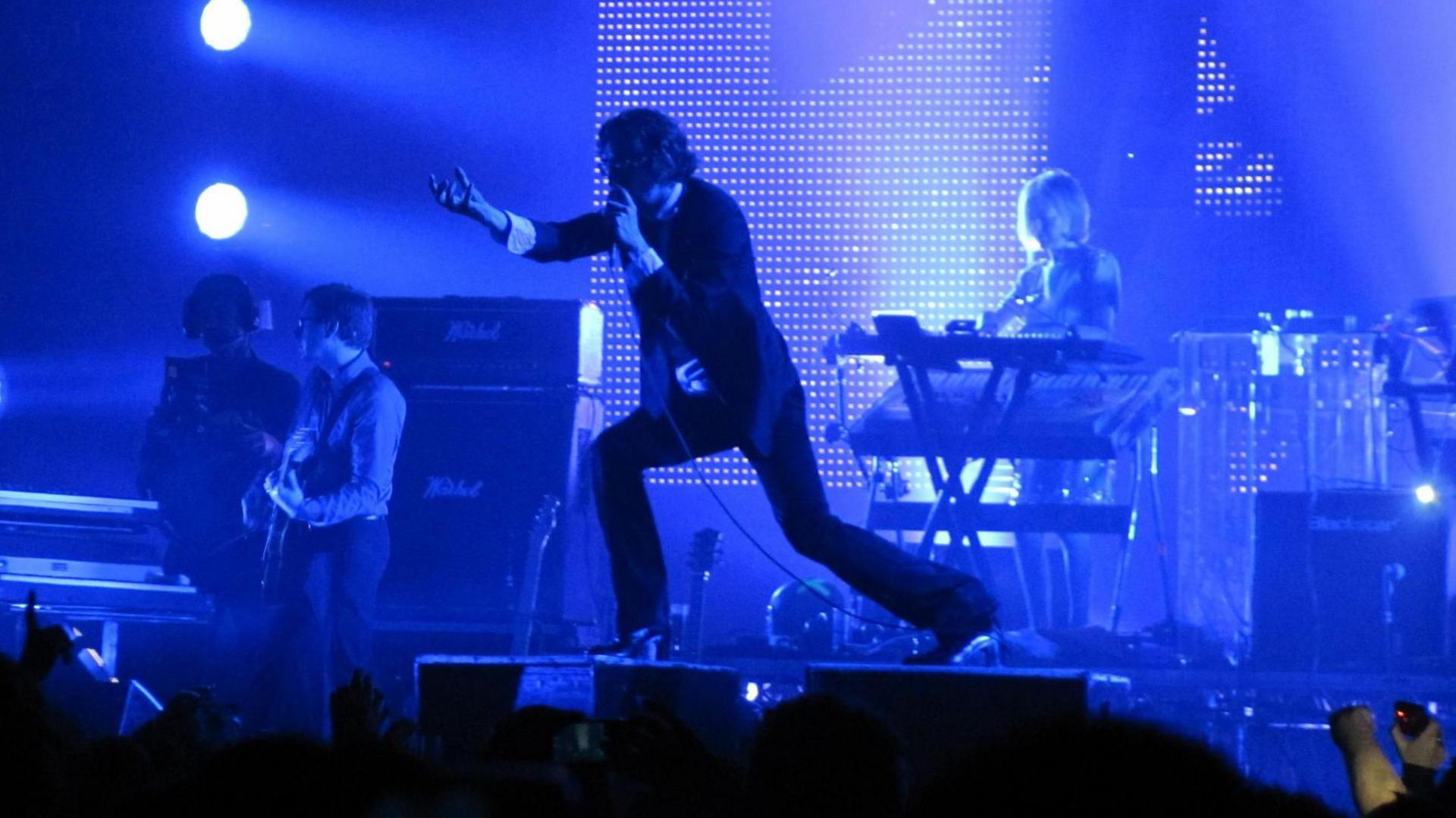 A lead singer on stage dancing whilst a keyboard player and guitarist perform behind him. A blue light covers all the performers.