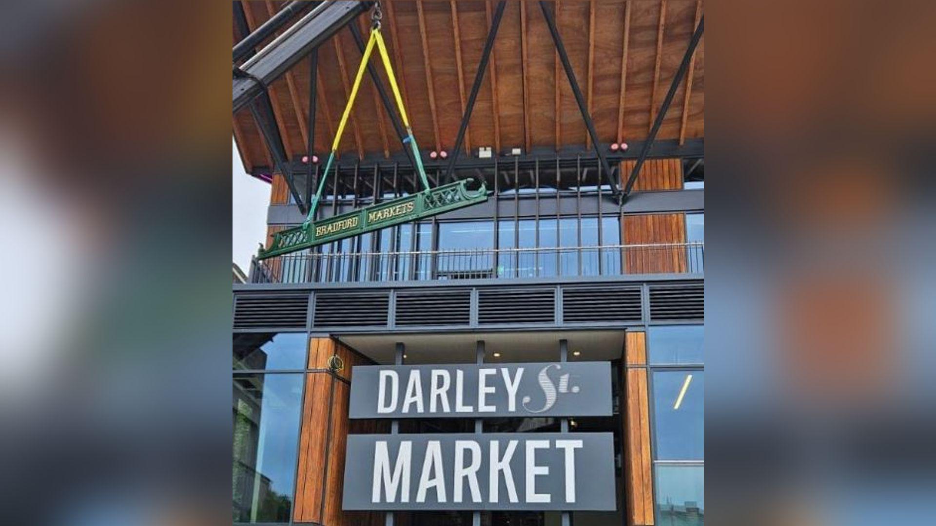 Darley Street Market building in Bradford with an iron sign reading Bradford Markets being winched by crane into position on the top floor of the three-storey building.
