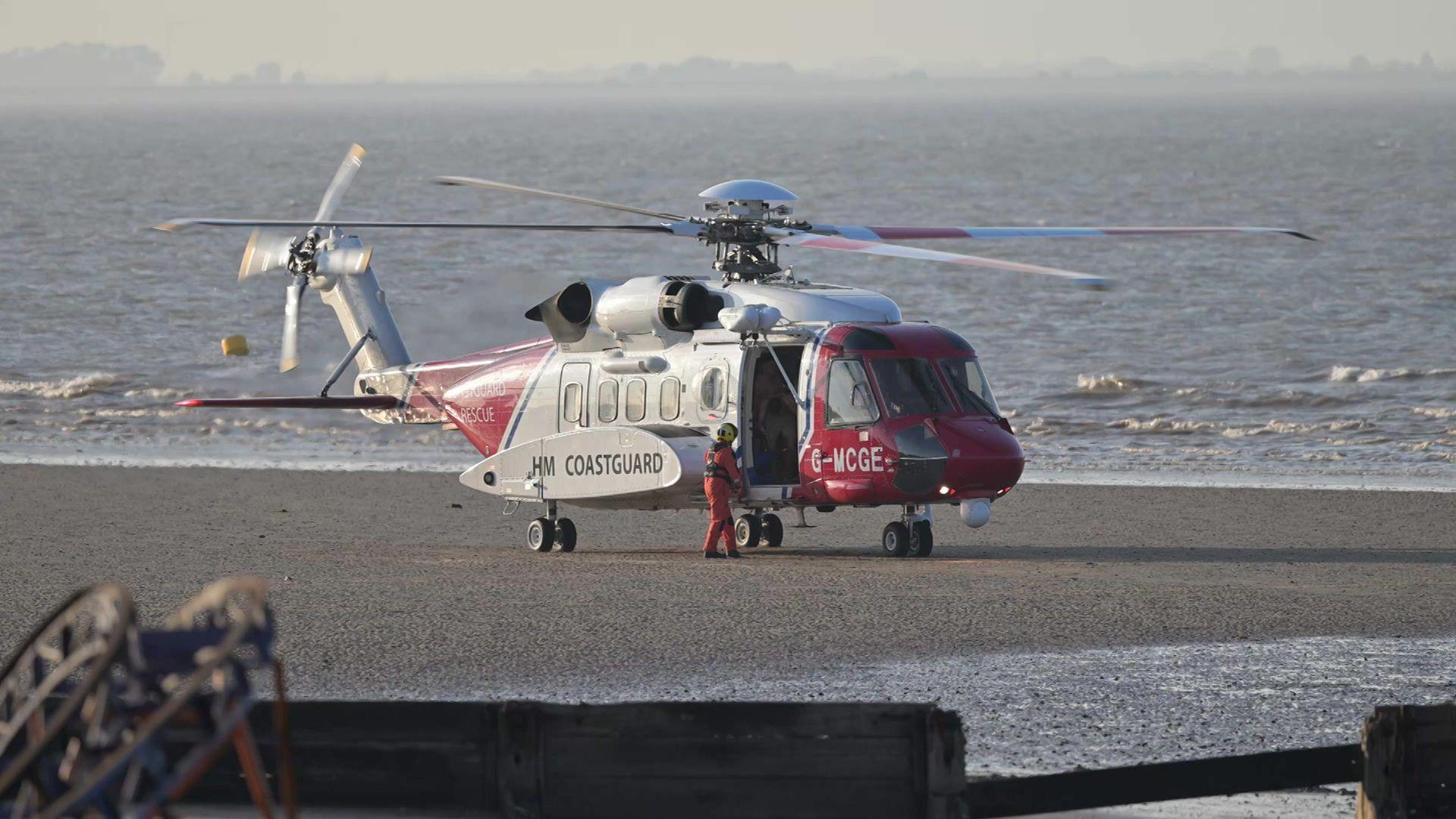 A red and white helicopter with the word "HM Coastguard" in black on the side. A crew member, wearing a yellow helmet and orange overalls, is standing on the sand next to the helicopter. Water is visible in the background.