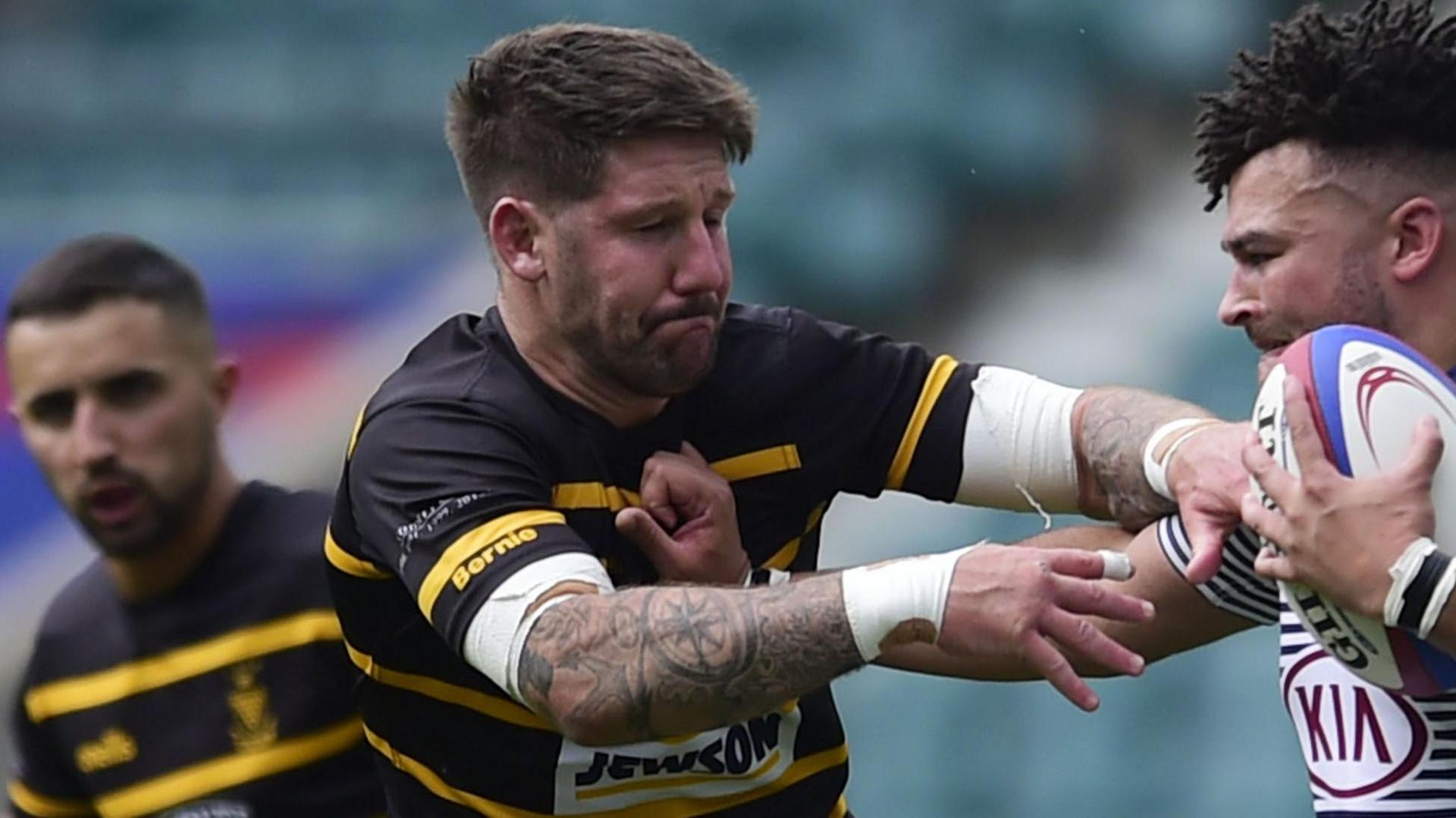 Matt Shepherd wearing a black and yellow-striped Cornwall shirt is in action against a rival player with a team-mate in the background.