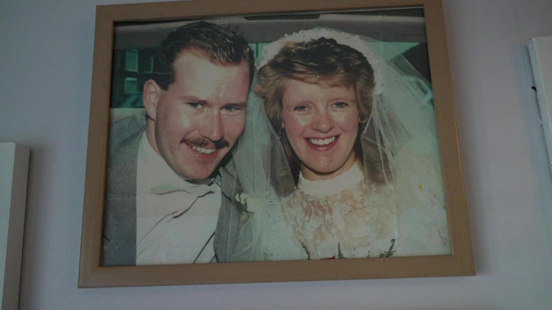 Barrie and Mo smile for the camera on their wedding day. She is wearing a white wedding dress while he is in a grey suit