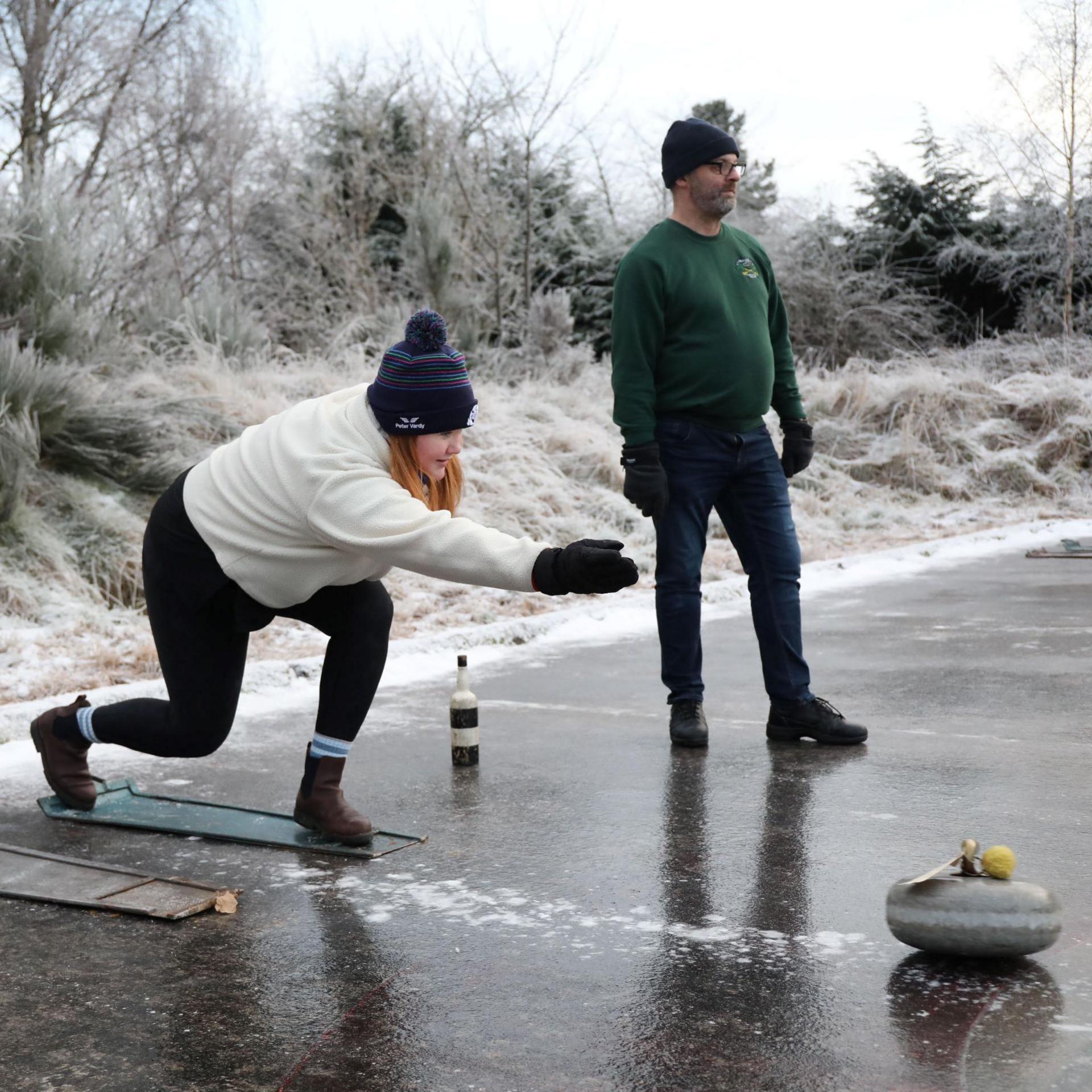 A curler bends down to release a stone down the rink while watched by a fellow curler. They are wearing woolly hats and gloves.