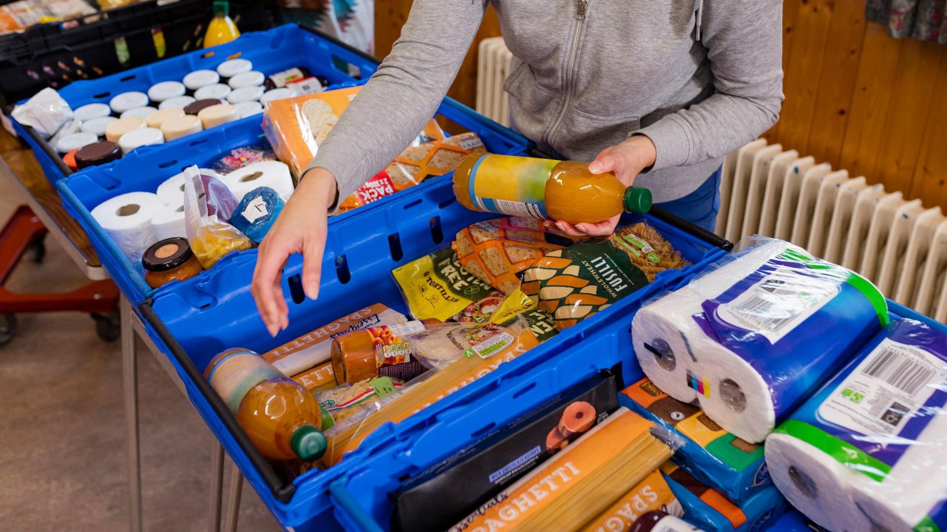 A line of blue plastic boxes with food and toilet roll in them. A person in a grey top is holding a bottle of squash and has another hand hovered over the boxes.