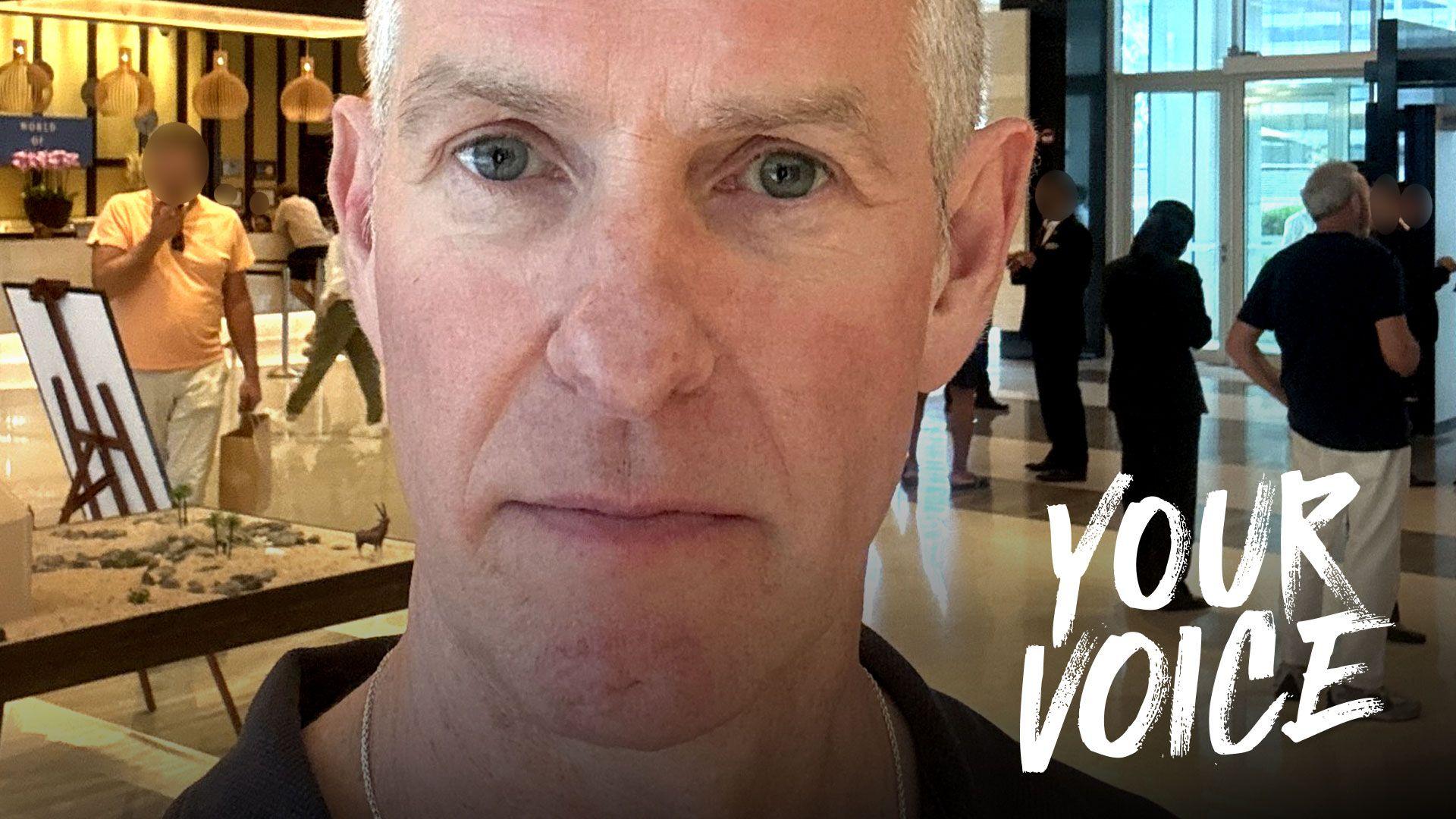 A man looks serious while standing in front of people milling about an airport shopping area.