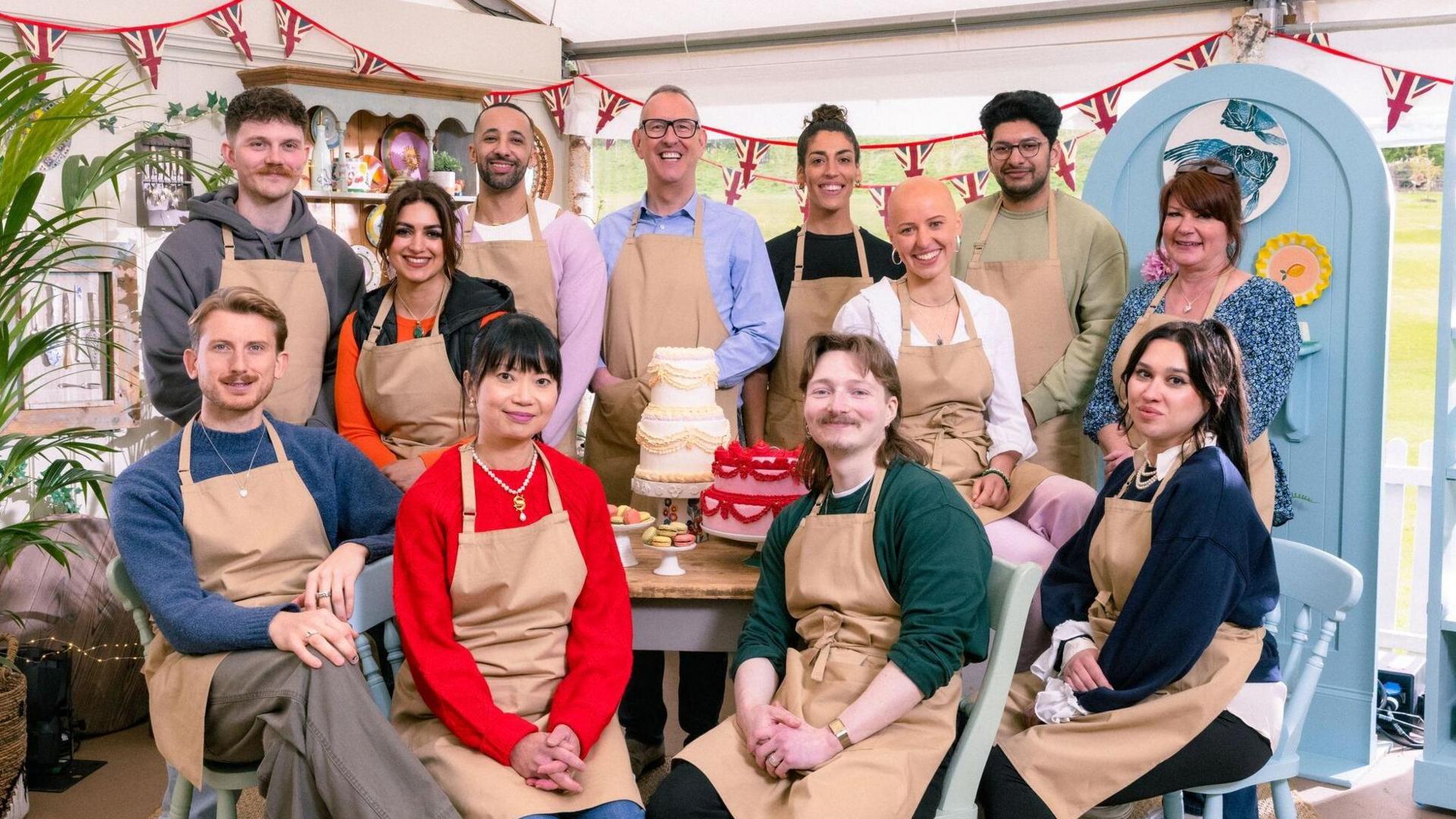 Twelve Bake Off contestants - six men and six women - sit and stand around a wooden table with cakes on it. They are all wearing tan-coloured aprons and are smiling at the camera. A white and a pink tiered cake sit in the middle of the table.