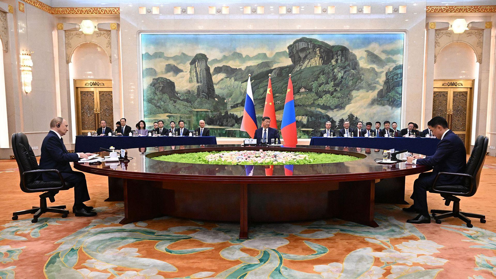 The three leaders sit at opposing ends of a huge circular wooden table with greenery and rocks in the middle of it, with a huge scenic landscape painting hanging on the back wall of the room with the three flags and delegates sitting at tables at the back.