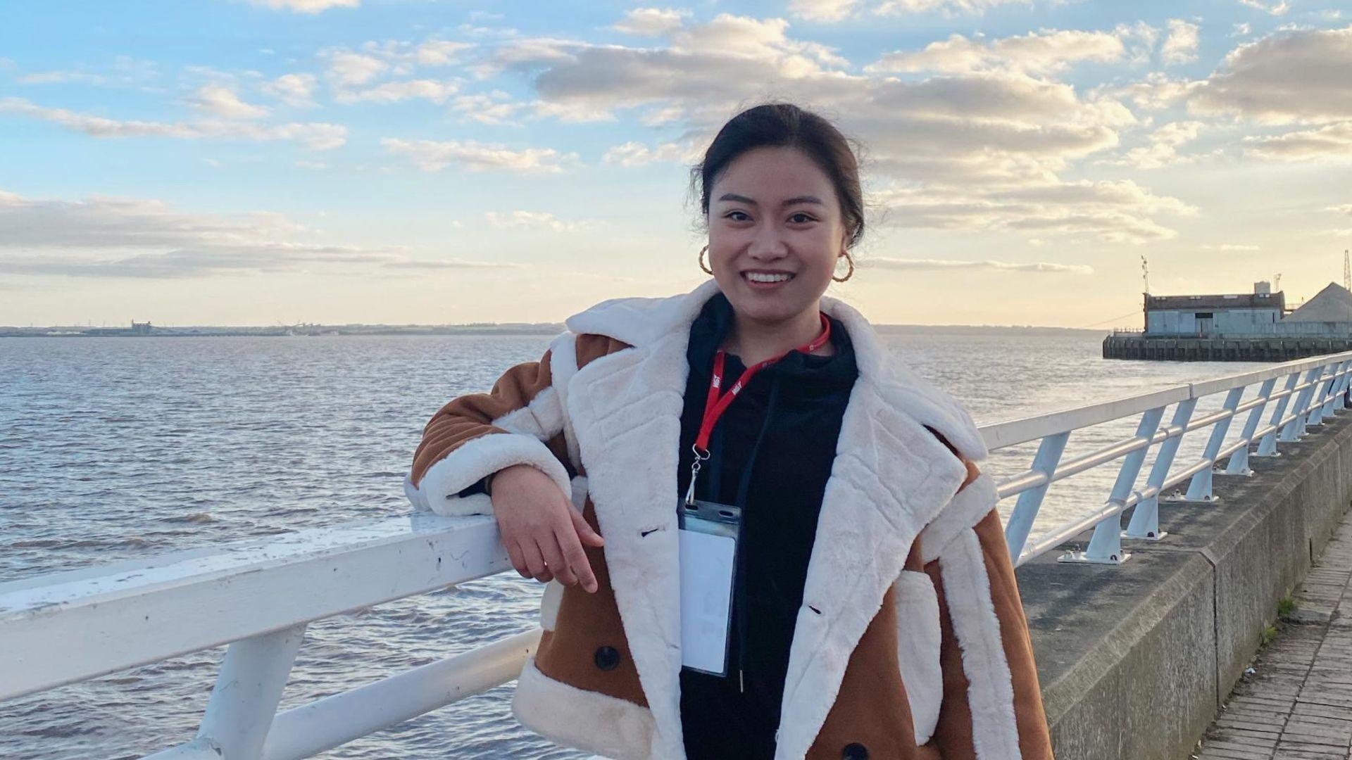 Yu, leaning against a white barrier on a pier. She smiles at the camera. She wears a brown jacket with white fur trim. She has a black fleece on, and a lanyard around her neck. The sky has a few clouds which are lit up by the sun setting.