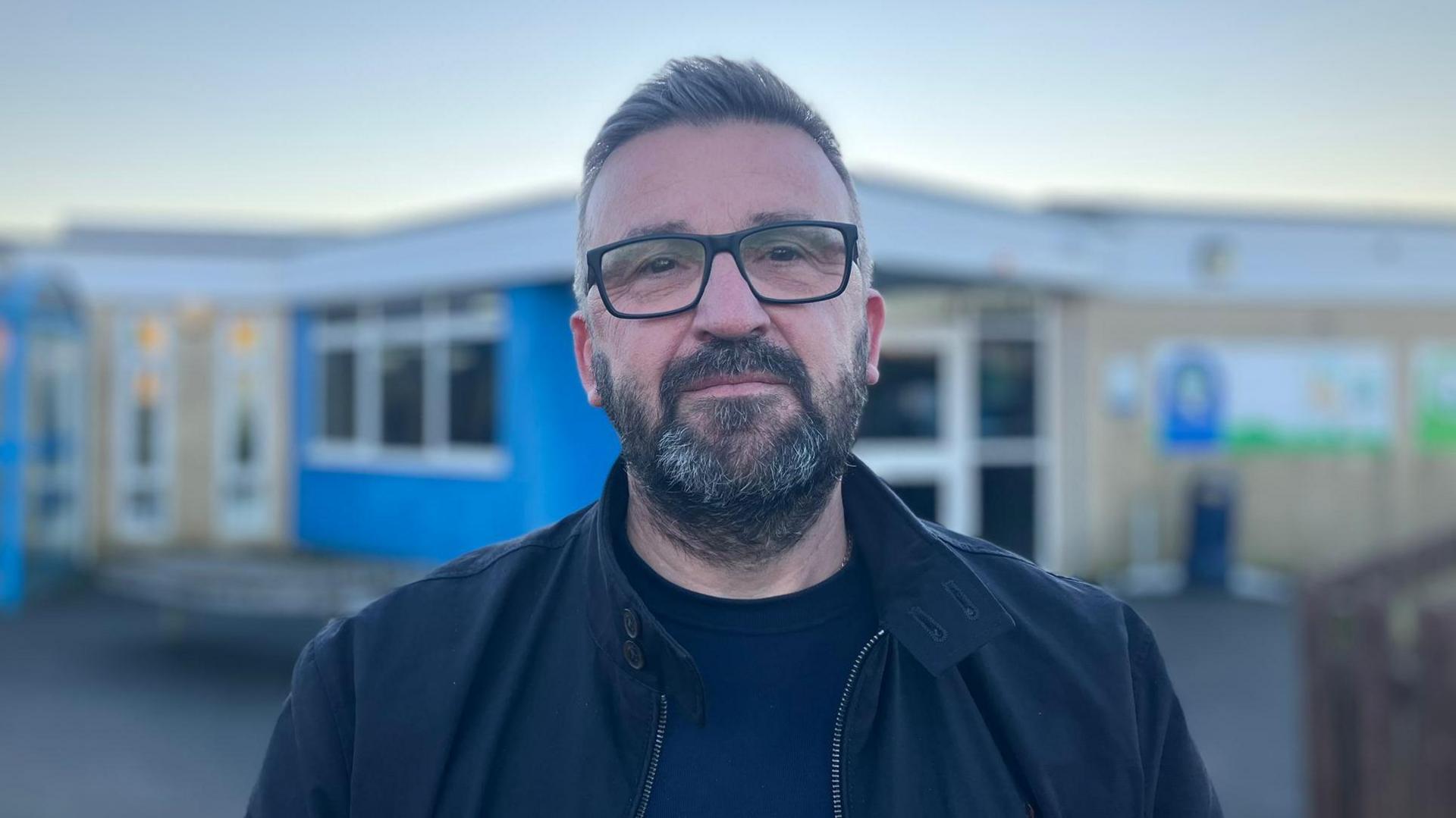 Nicholas Humphrey looks at the camera. He is wearing black glasses, navy top and black coat. The school is in the background.