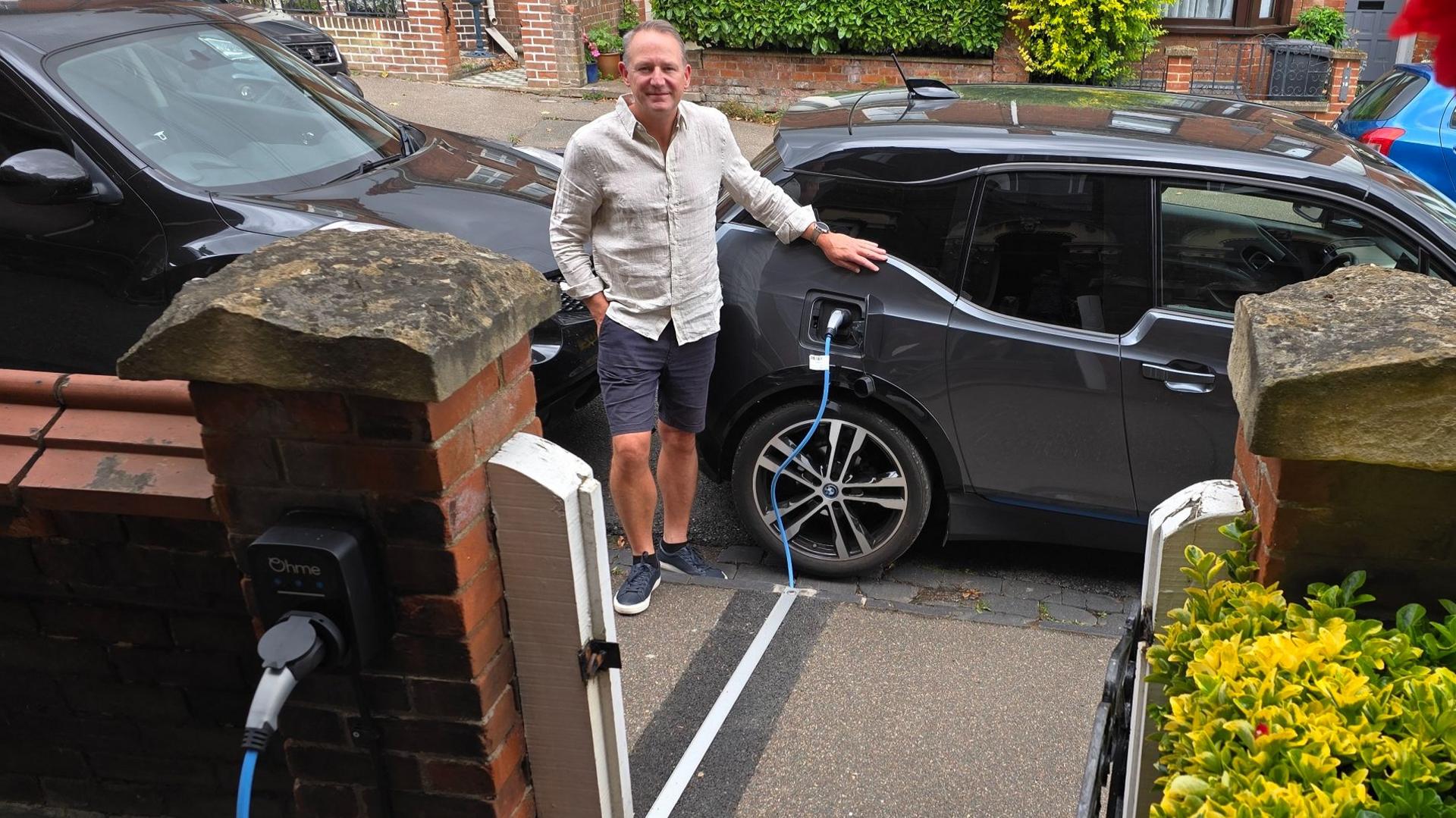 Chris stands on the pavement next to his black electric car, he wears navy shorts and a beige long sleeved shirt. The car is parked on the street, and the blue charging cable runs into a gully through the pavement, covered by a narrow metal strip. It connects to the wall of a terraced house which has red brick walls.