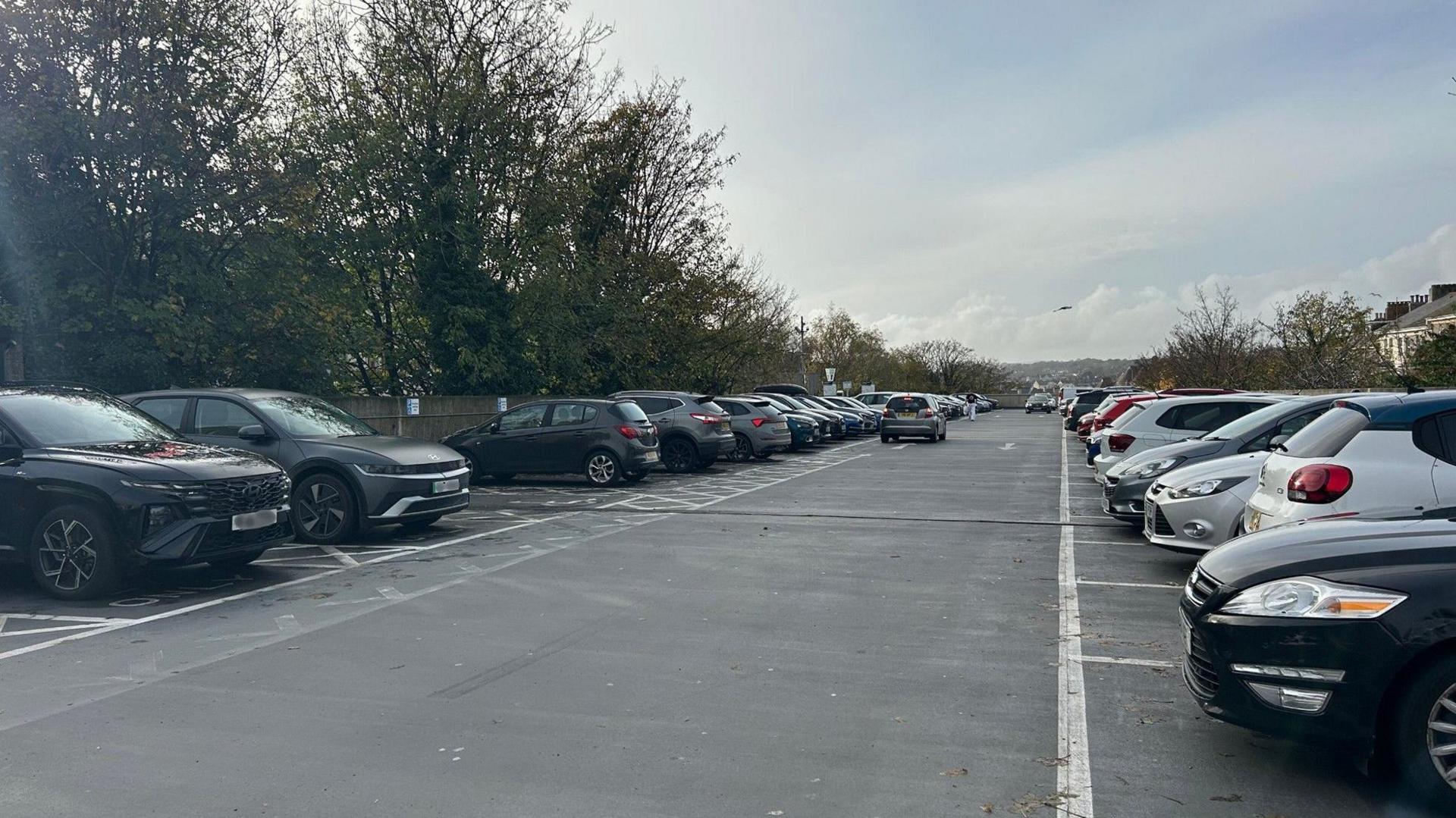 A number of parked cars in the Napier Terrace car park in the Mutley area of Plymouth. The car park is near enough full with lots of cars parked. There are designated disabled bays visible with lots of trees along the side of the car park. 