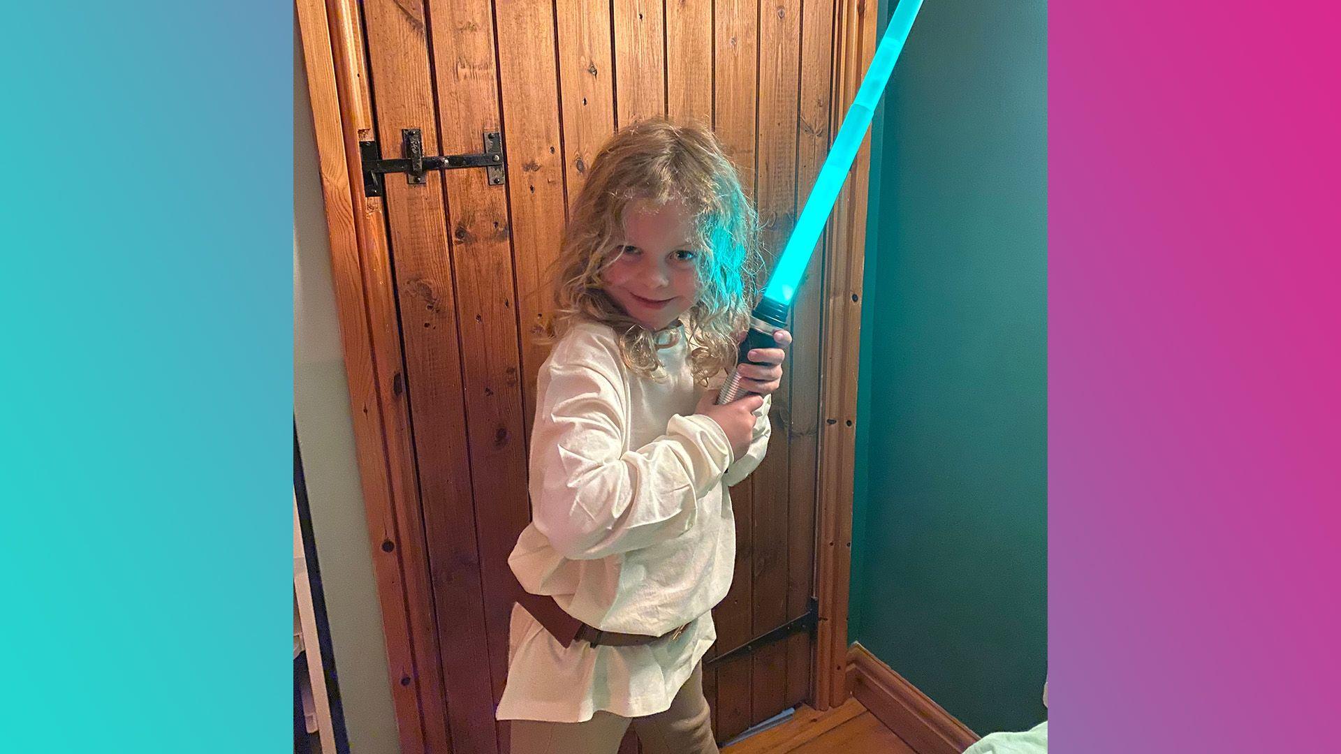 Lyra is wearing a sand coloured top, light brown trousers and a brown belt. They have shoulder length blonder hair and are holding a blue lightsaber