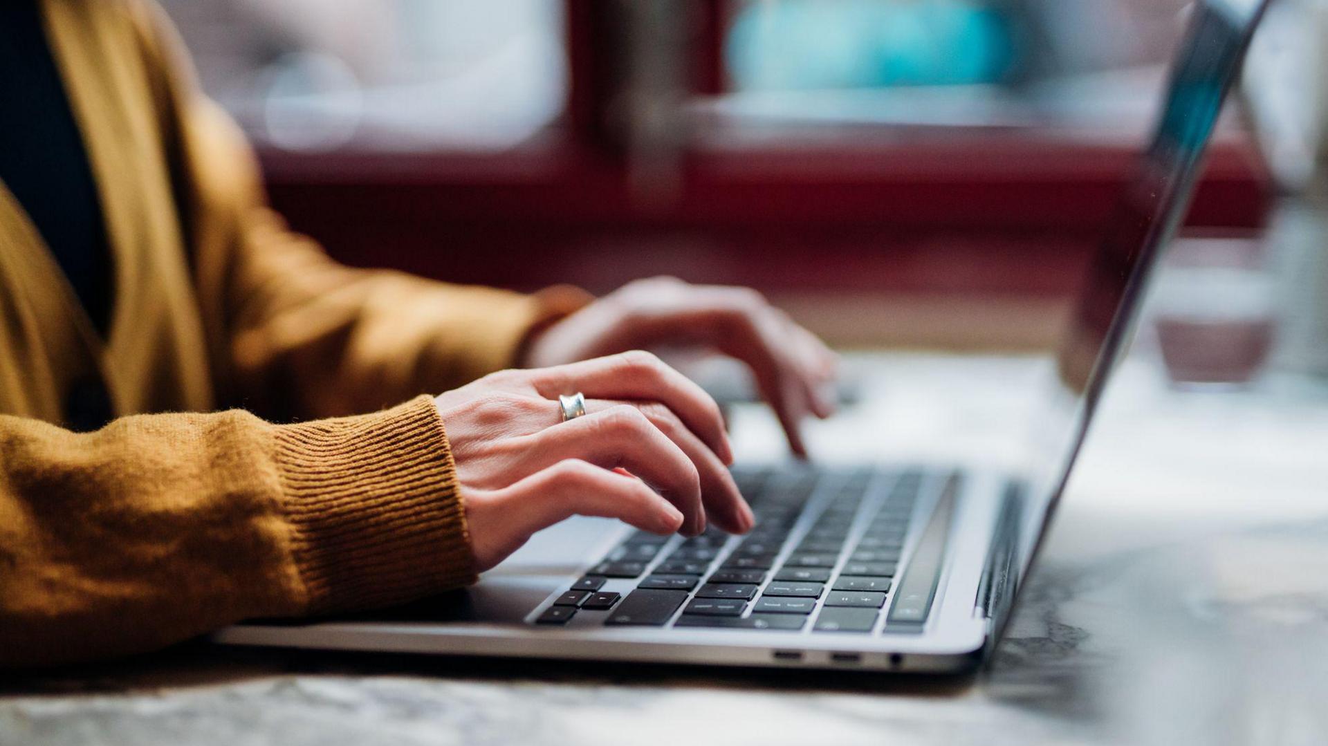 A woman - who is anonymous in the image and only her arms and hands can be seen - types on a laptop