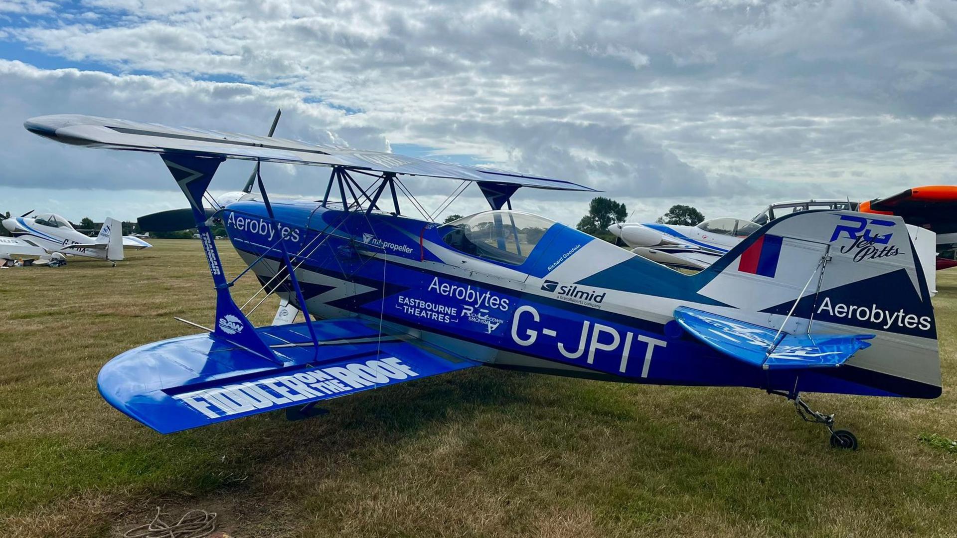 The blue and white G-JPIT aircraft parked alongside other small planes in a field. It has wings above and below its main body.