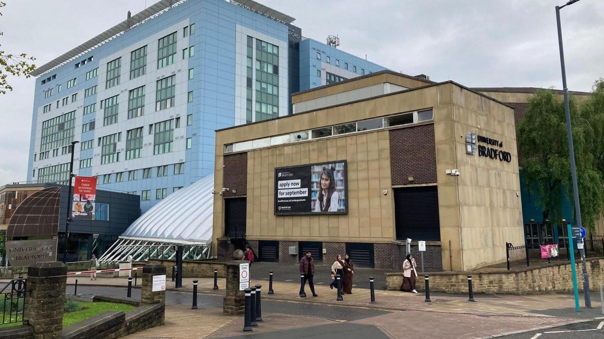 Two buildings located on a road side with people walking past. One of the buildings is several storeys high. The other building is smaller and has a sign on the side saying University of Bradford