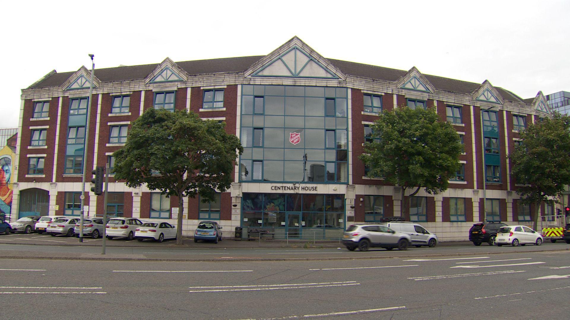 The outside of a large building. The centre is a large window the full length of the building with many windows across it. The frames are blue and white and it is red brick. A number of cars, mostly white and silver, are parked outside facing towards the building. A Salvation Army logo can be seen on one of the windows in the centre of the building, above a sign reading "Centenary House"