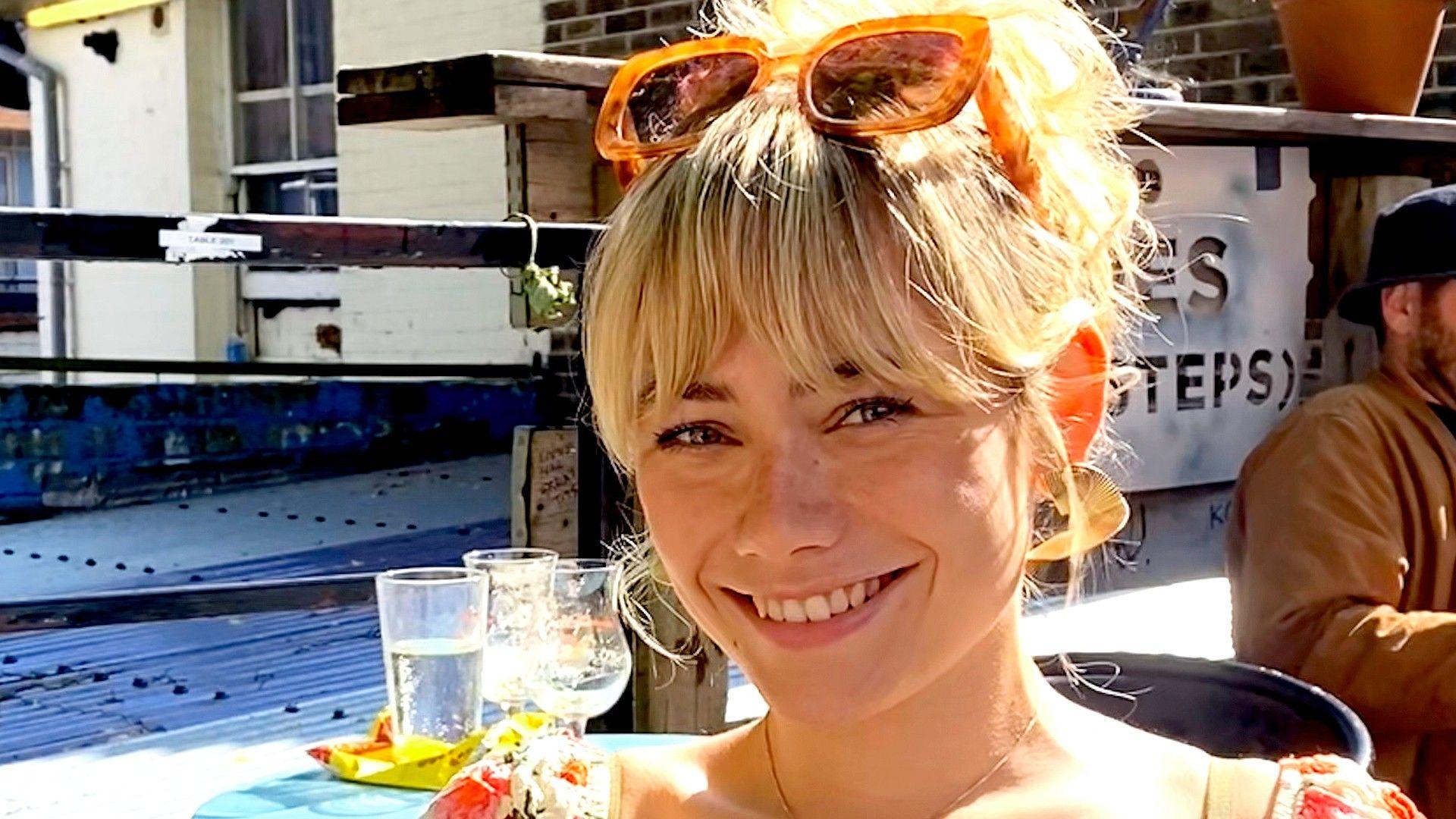 Izy has her blonde hair tied up with sunglasses on top of her head. She sits smiling in an outdoor location with buildings in the background.