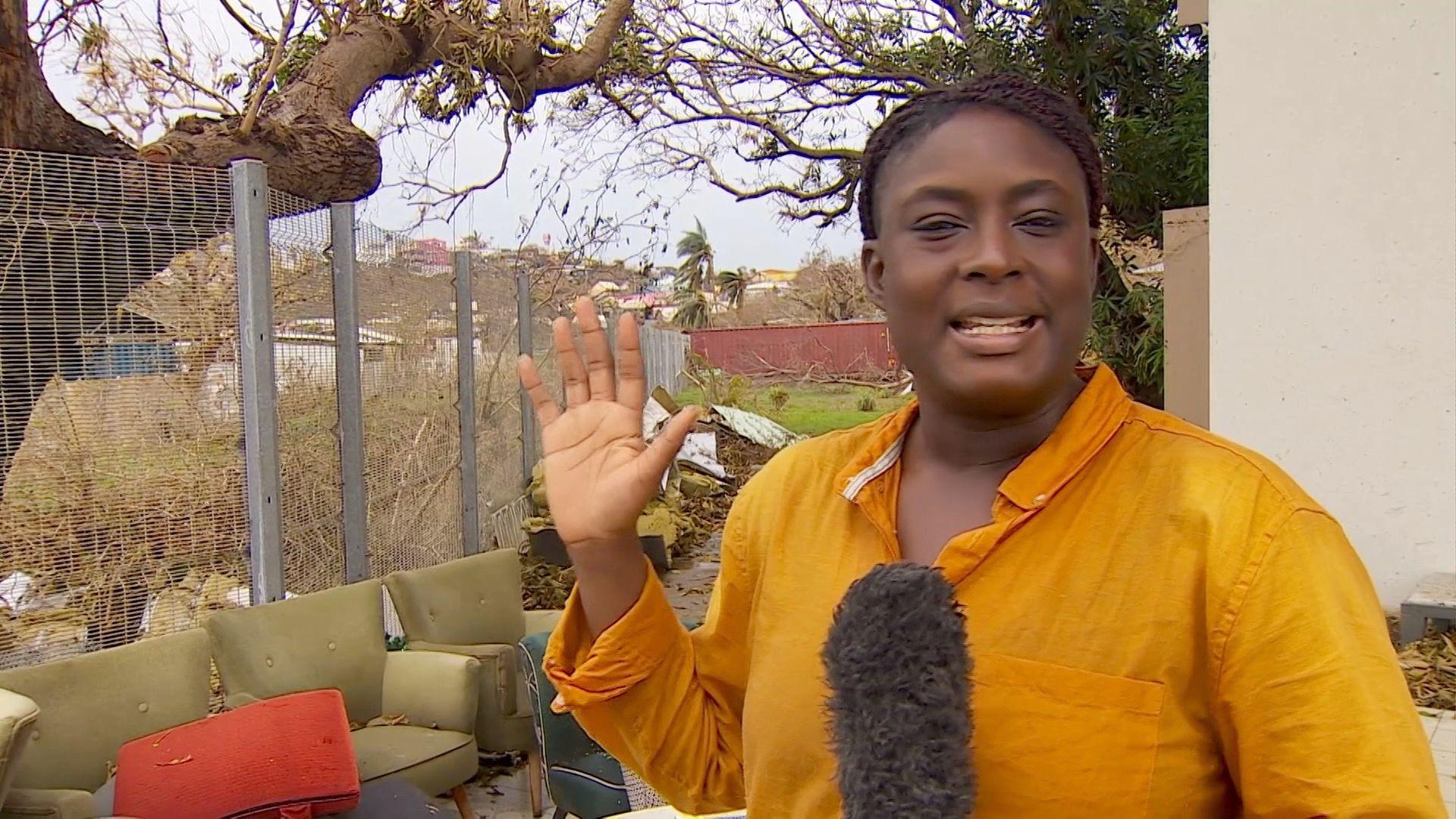 Mayeni Jones in a yellow shirt talking into a microphone against a backdrop of scattered furniture and trees stripped of their leaves