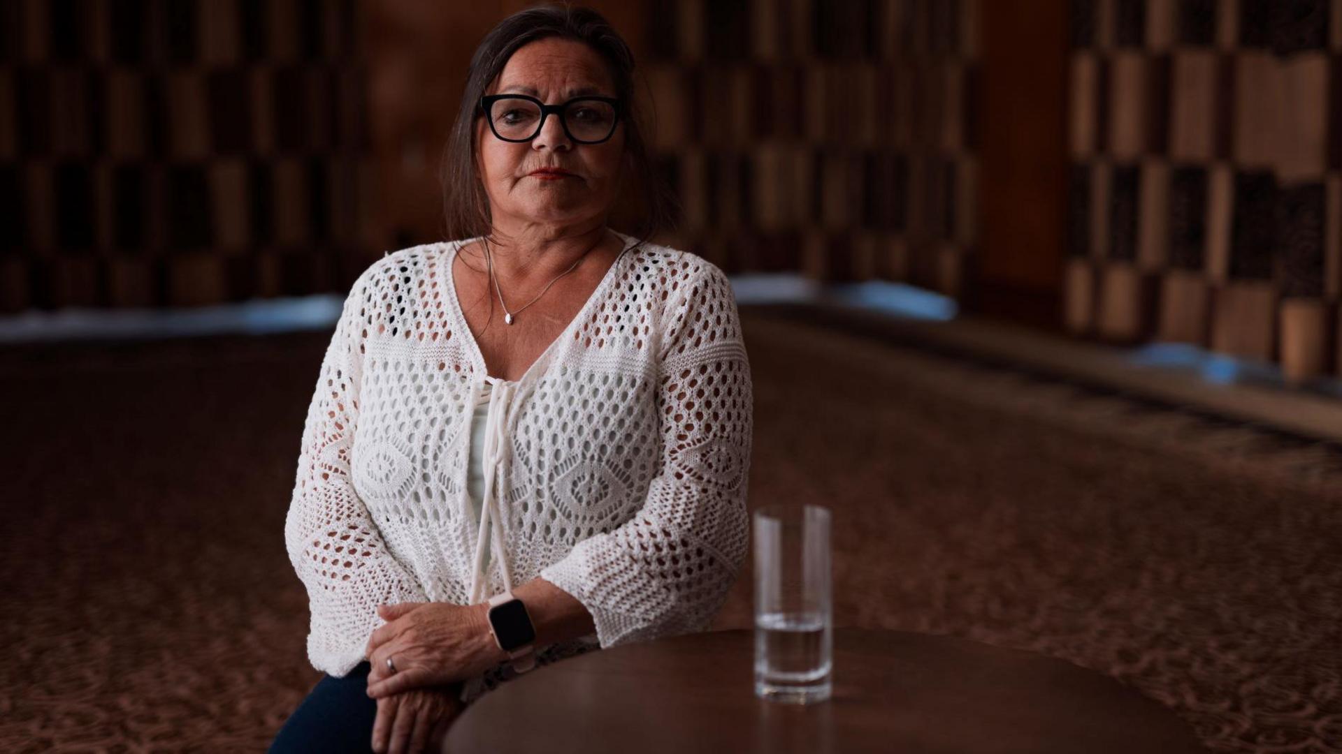 A photo of Fee against a dark background. She has dark hair and glasses. She is wearing a white crocheted cardigan
