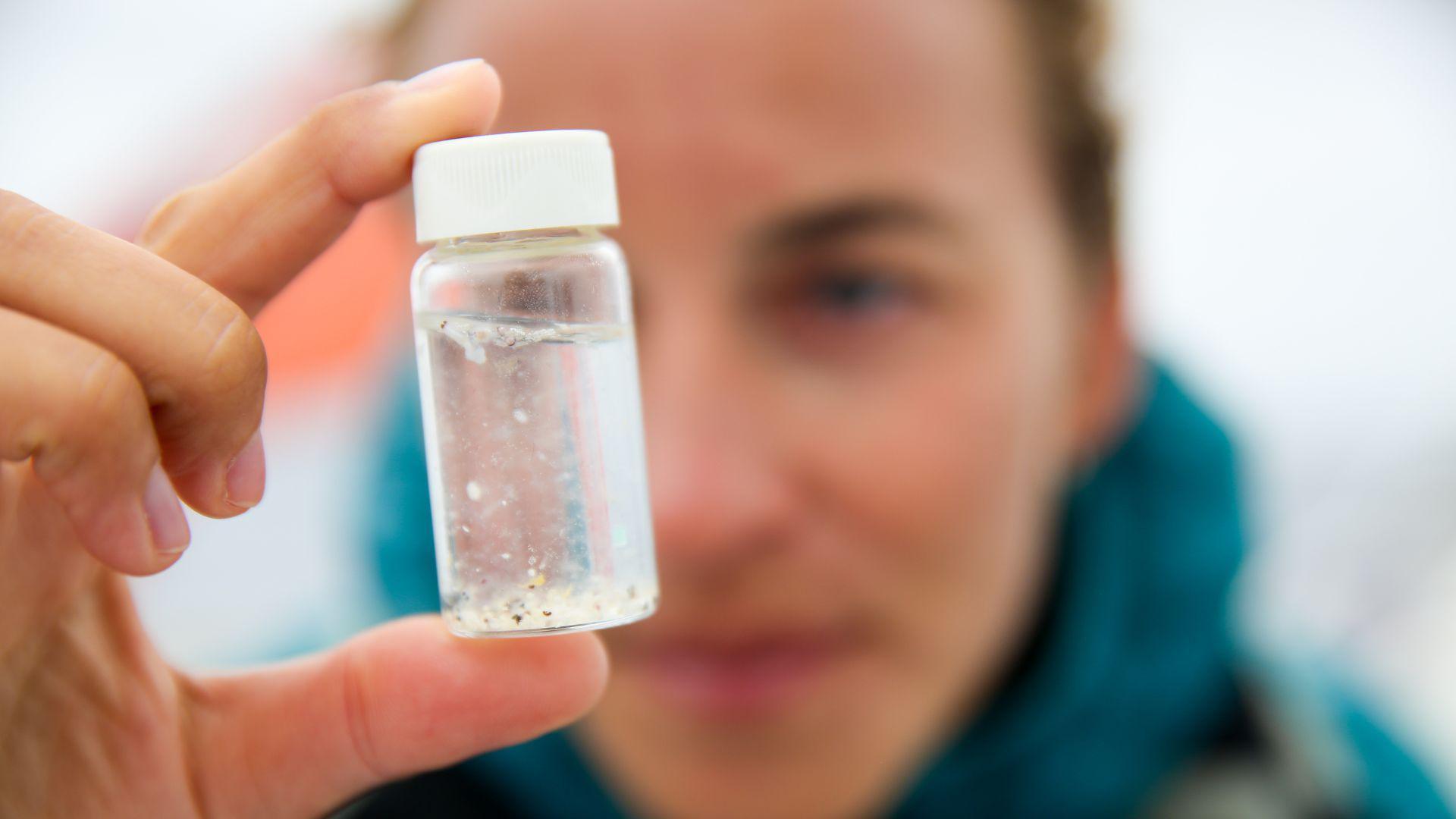 A person holding a small sample of water in front of their face. There are small pieces of plastic in the water.