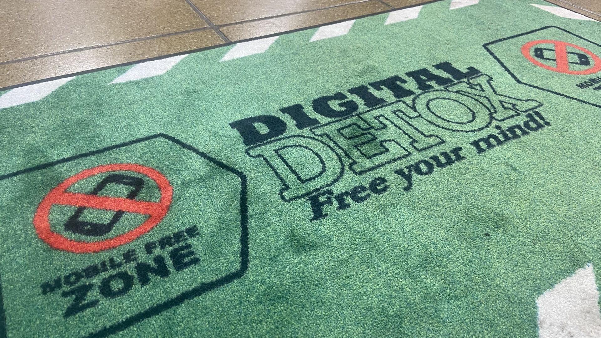 A green door mat with large words saying "digital detox" and a no entry sign style logo covering up an image of a mobile phone.
