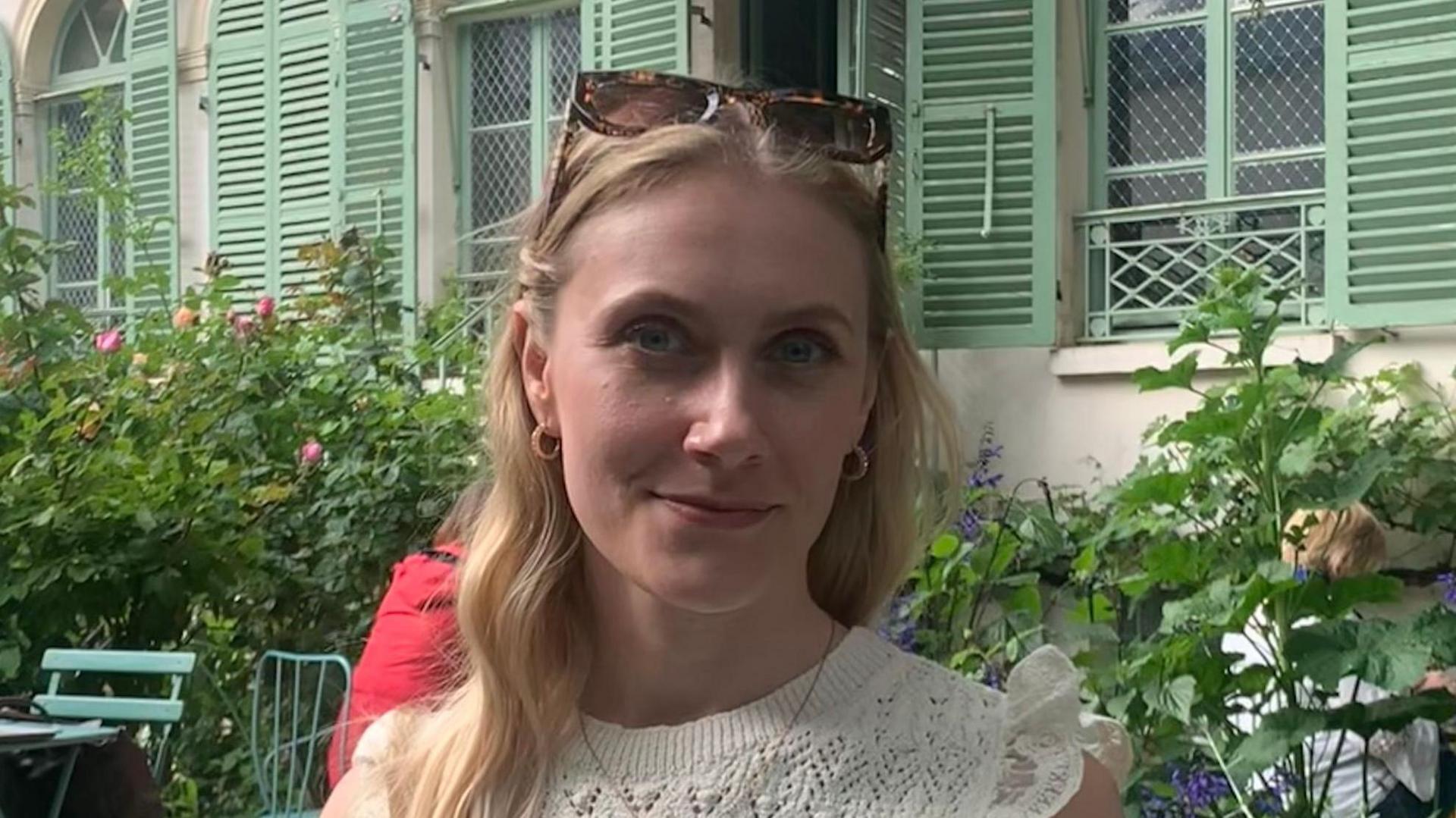 Lili Vachon wears sunglasses on her head as she poses for photo in a garden cafe with plants and a building behind that has green window shutters. She has long blonde hair and wears a white top.