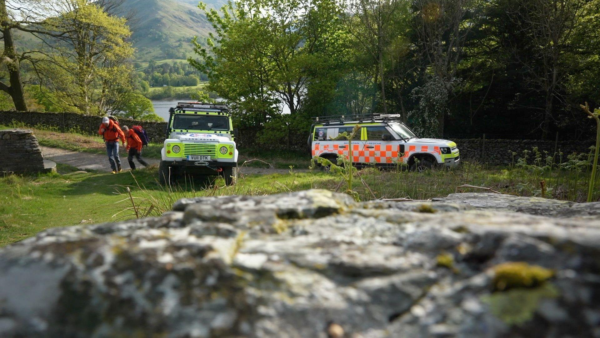 Two white Land Rovers with orange and green liveries parked next to a road. Two people in orange coats are starting to walk away form the vehicles, with a lake and trees behind them