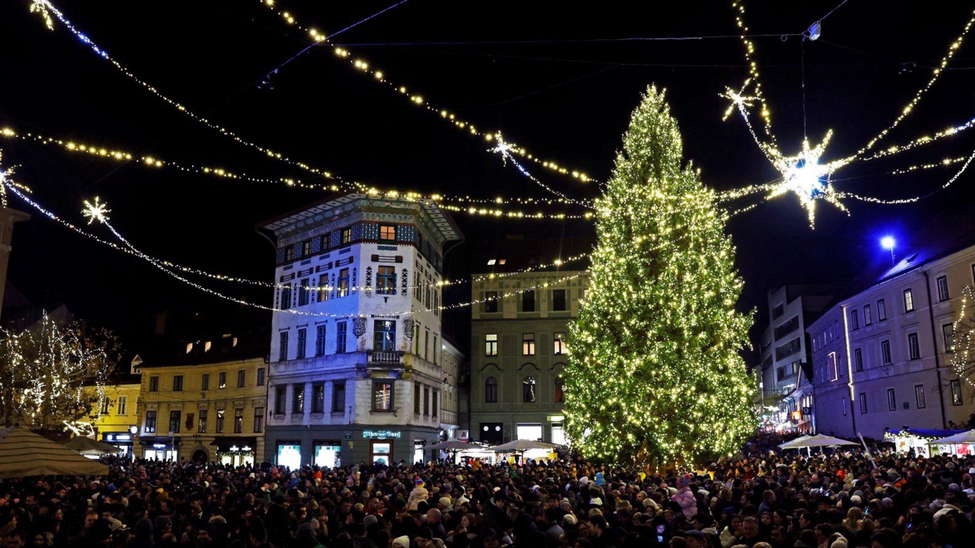 lights hang across a square with lots of people gathered around a tall christmas tree covered in warm white lights