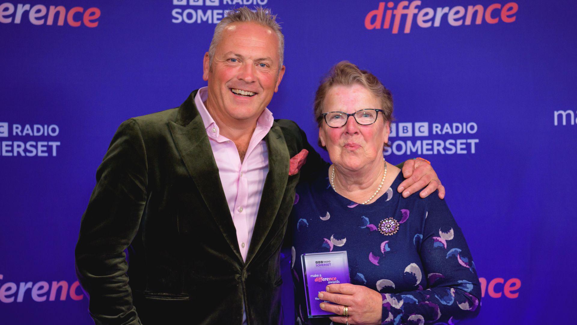TV presenter Jules Hudson wearing green velvet suit jacket stands to the left of lady wearing glasses and purple top with trophy in hand