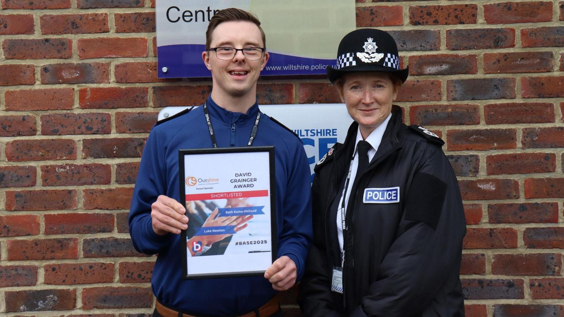 Luke Newton standing next to a female police officer. He has short dark hair, glasses and is wearing a blue shirt. He is holding a certificate with his name on that says "shortlisted". The female officer next to him is wearing a white shirt, black tie, a black police jacket and a police hat. Both of them are looking at the camera and smiling.