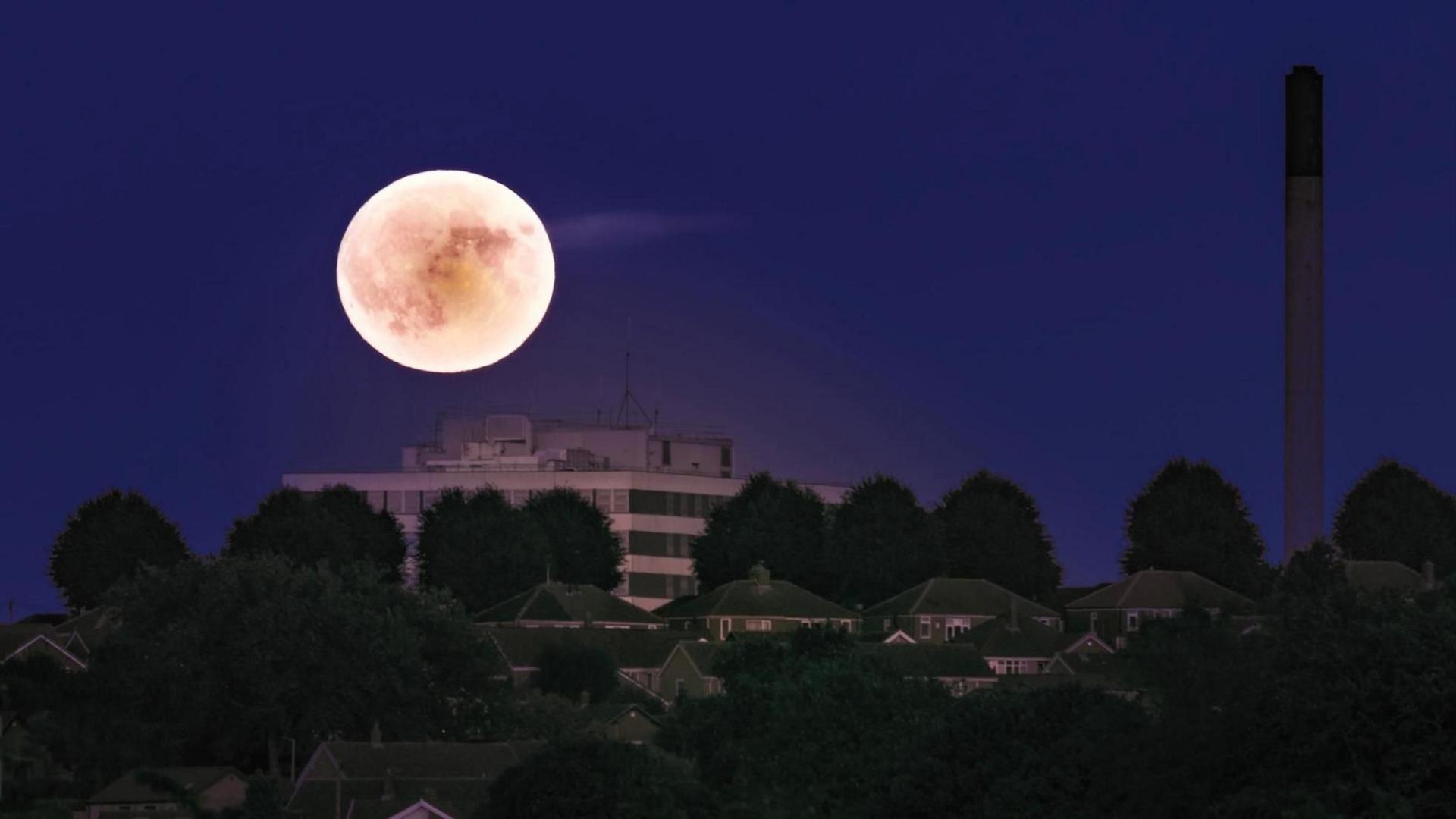 A glowing supermoon above a rectangular mult-storey building. Four houses can be seen in a row in front of it and a factory chimney towers to the right.