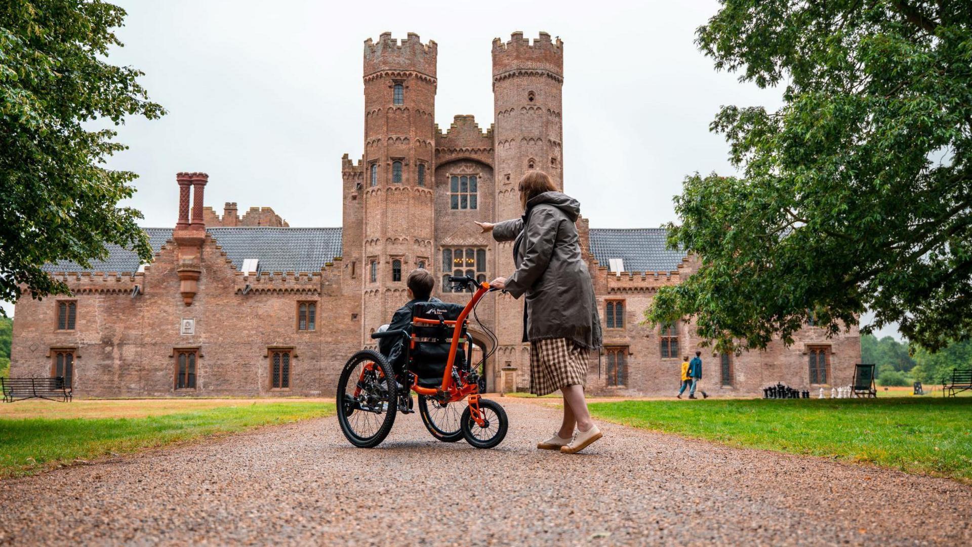 A woman with her hand on a powered wheelchair with a young boy sitting in it. They are visiting Oxburgh Estate - and can be seen outside Oxburgh Hall, a moated medieval house.