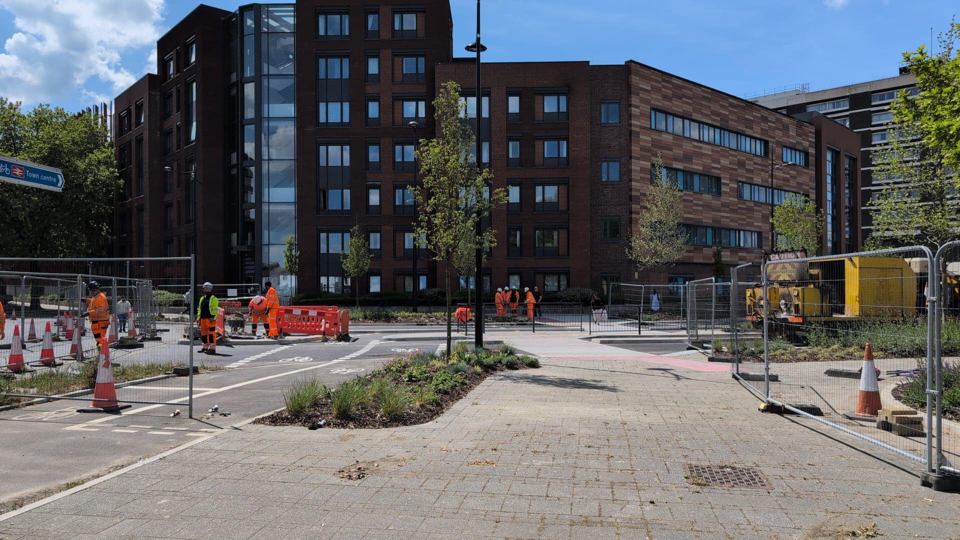 A foot way leads towards a multi-storey office building. The road next to the path is blocked off for road works, with people in orange high-vis working on it.