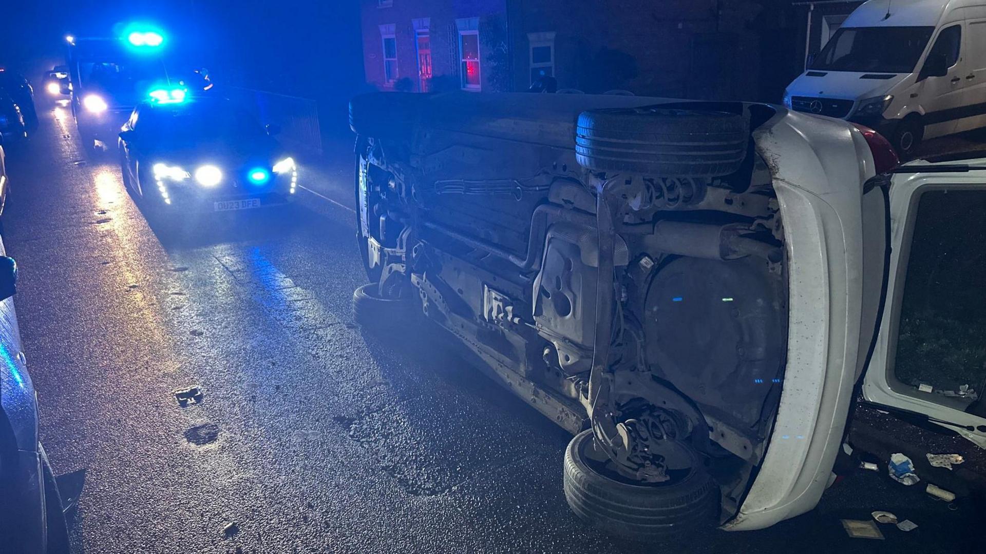 A white car on its side after a crash in a residential road. There are both a police car and an ambulance behind it with blue lights on. It is dark.