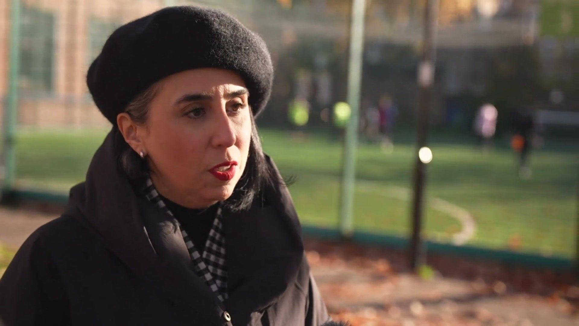 Eva Guerra in front of the football pitches. She wears a black beret and a black coat. She has red lipstick.