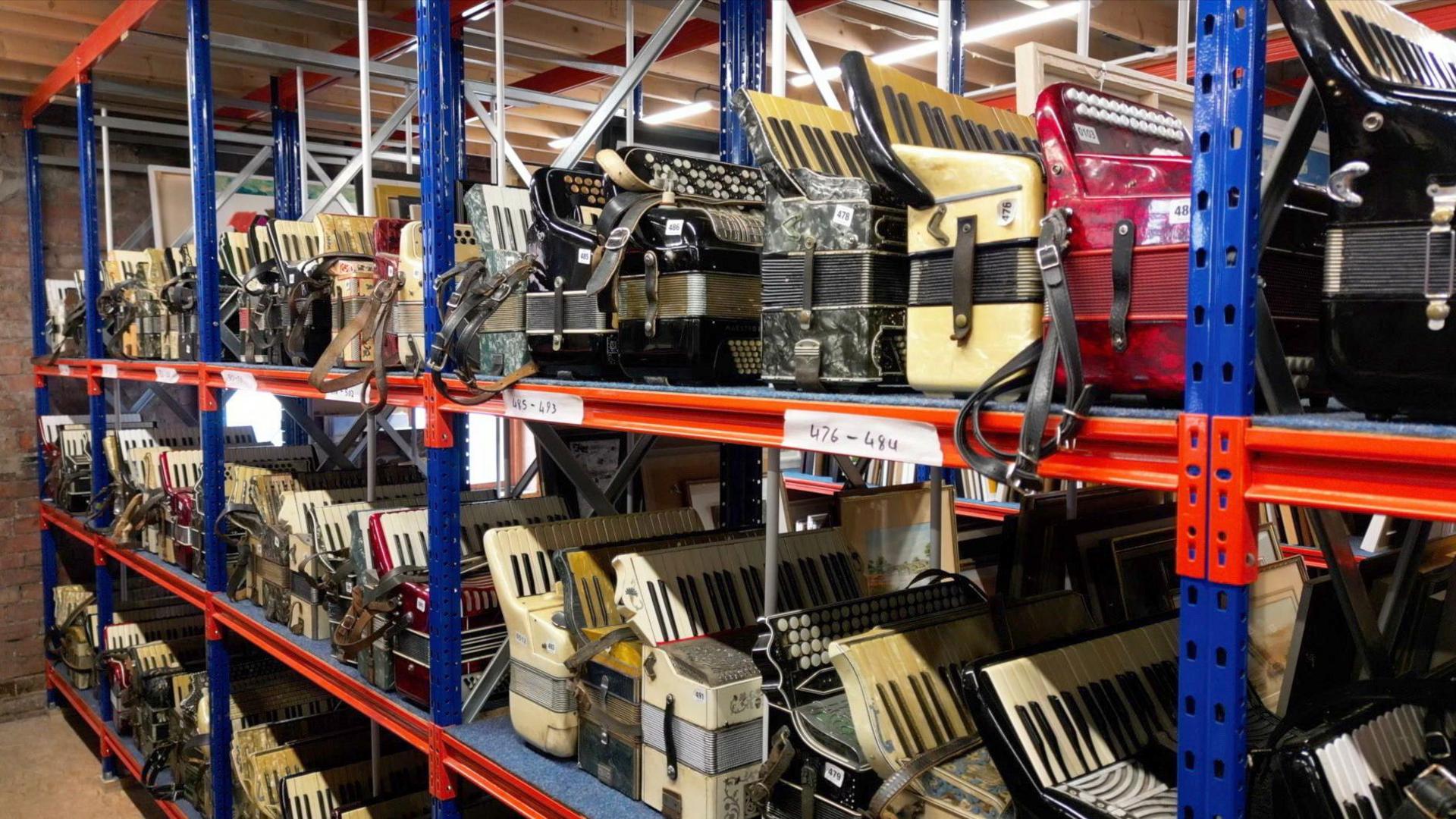 Row of accordions stored on large metal shelves in a warehouse. The accordions are of different shapes and sizes and the shelves are painted red and blue.