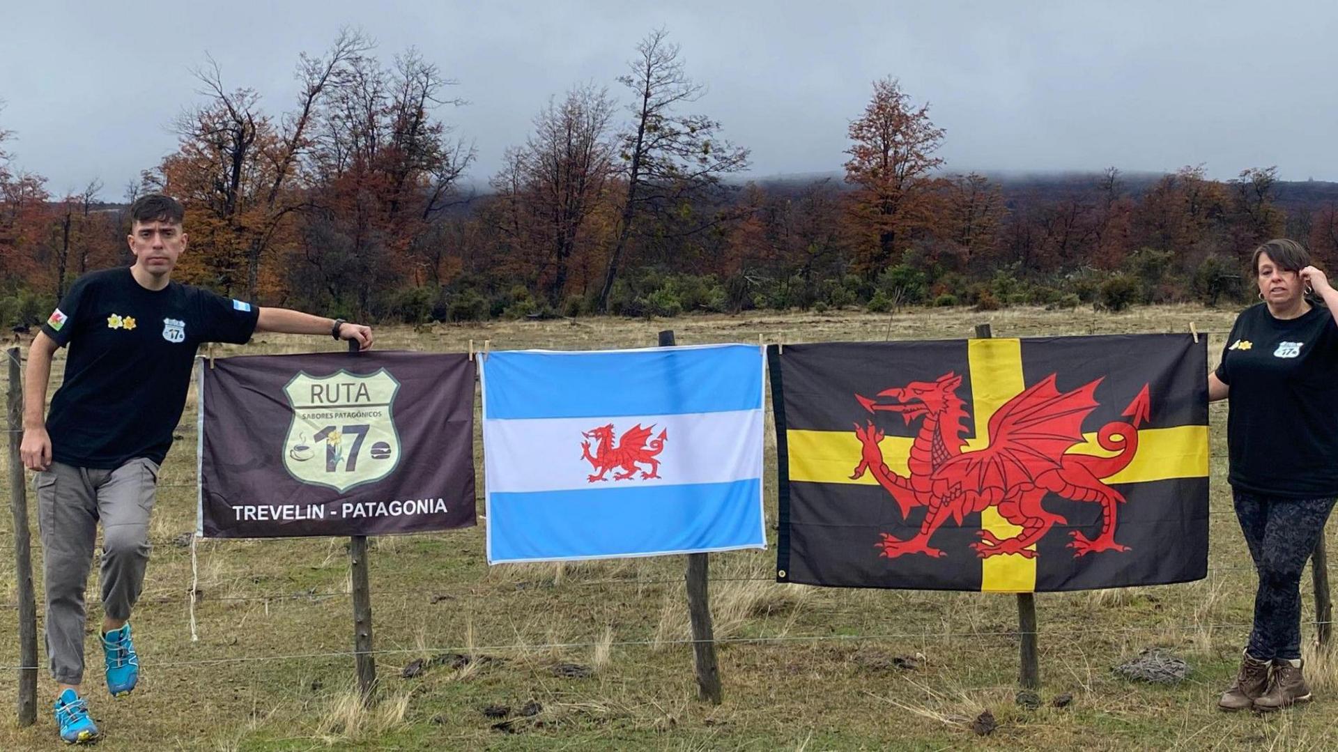 David and Peace stand either side of three flags. One has the sign for a route 17, the other an Argentine flag with the Welsh dragon and the third is a Black flag with a yellow cross and a large red dragon.