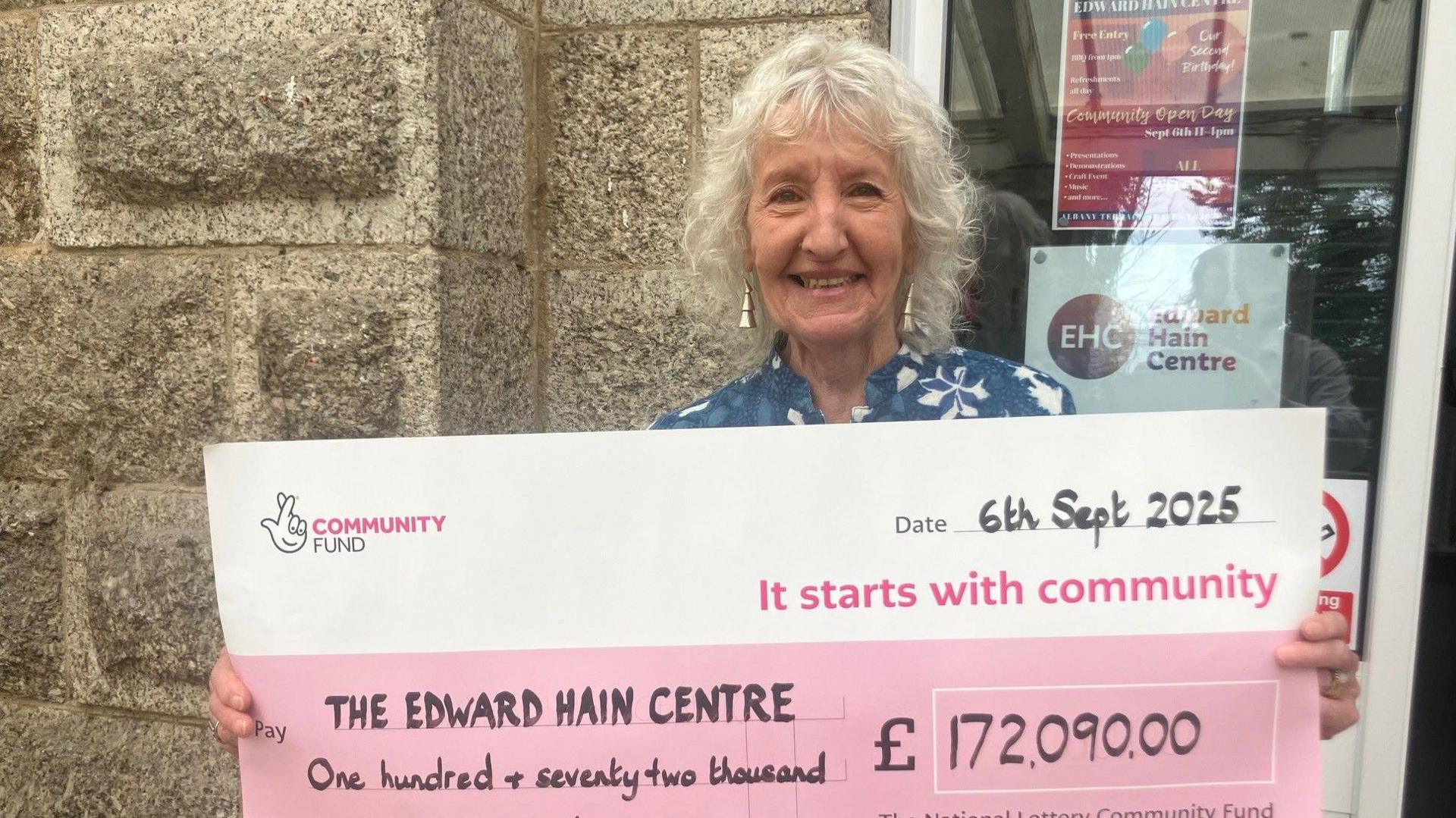 A woman stands in front of a granite building - she holds a giant cheque for £172090.00 from the National Lottery. She is smiling 