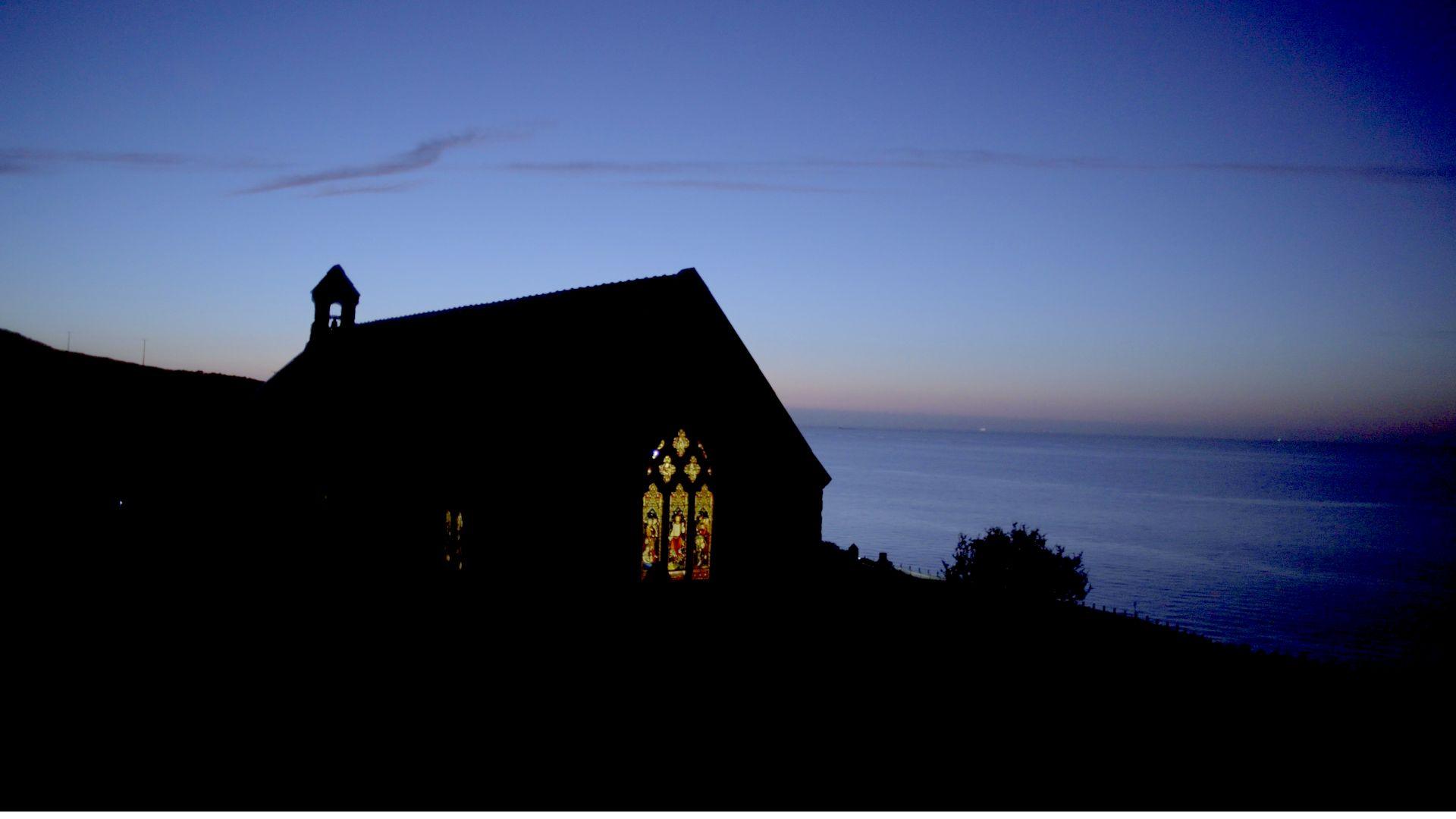 Llun silhouette o Eglwys Sant Tudno yn y nos. Y mor yn y cefndir ac ychydig o fachlud haul y tu ol i'r Eglwys.