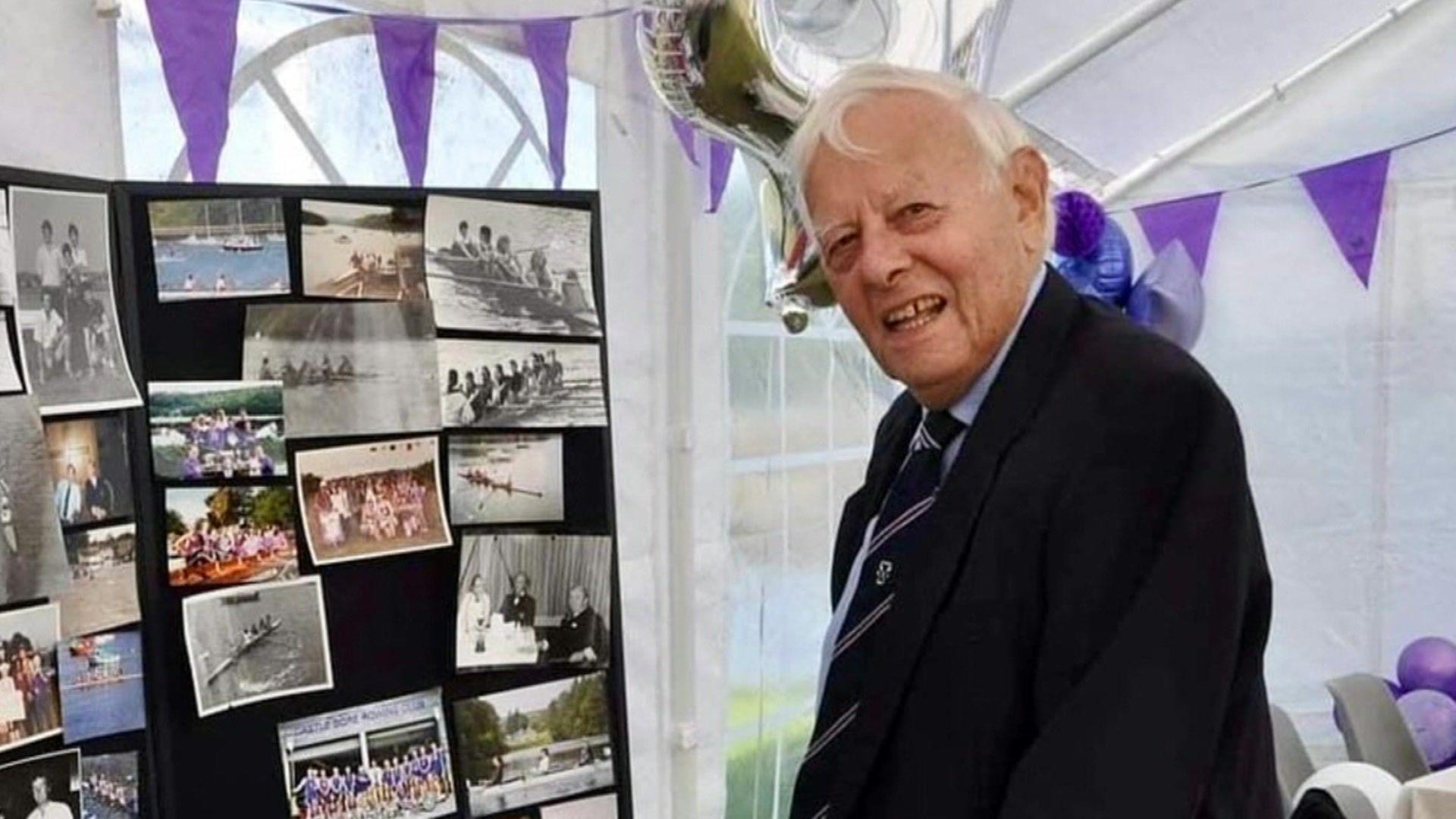 Morgan Bolding is seen in front of a board showing various rowing photos. He is in a marquee with purple bunting.