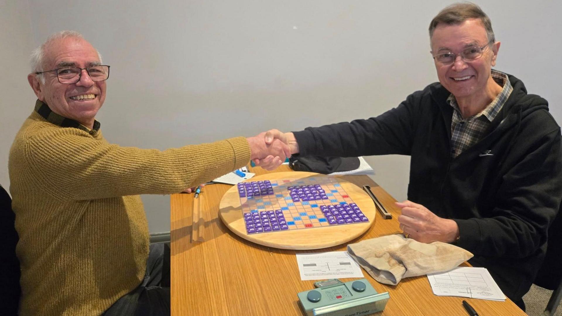 The two Bobs shake hands over a Scrabble board. Bob Lynn has short grey hair and wears glasses and a yellow jumper. Bob Linn has grey/brown hair and wears glasses and a black jacket with a green chequered collared shirt underneath.