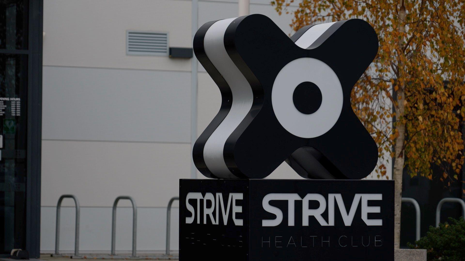 A black sign in the shape of an 'x', with a white circle in the middle. Beneath the shape, it says 'Strive' in white capital letters. Behind the sign is a tree with orange leaves and a series of bicycle racks.