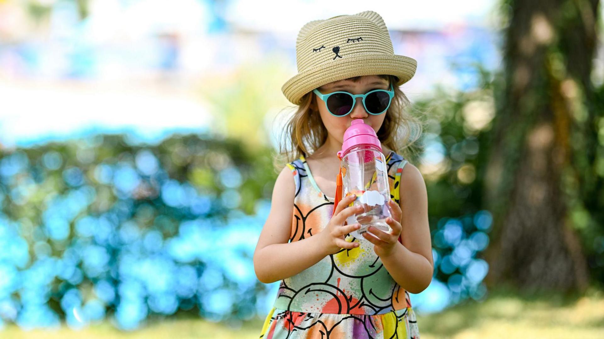a little girl wearing a hat, sunglasses and drinking water