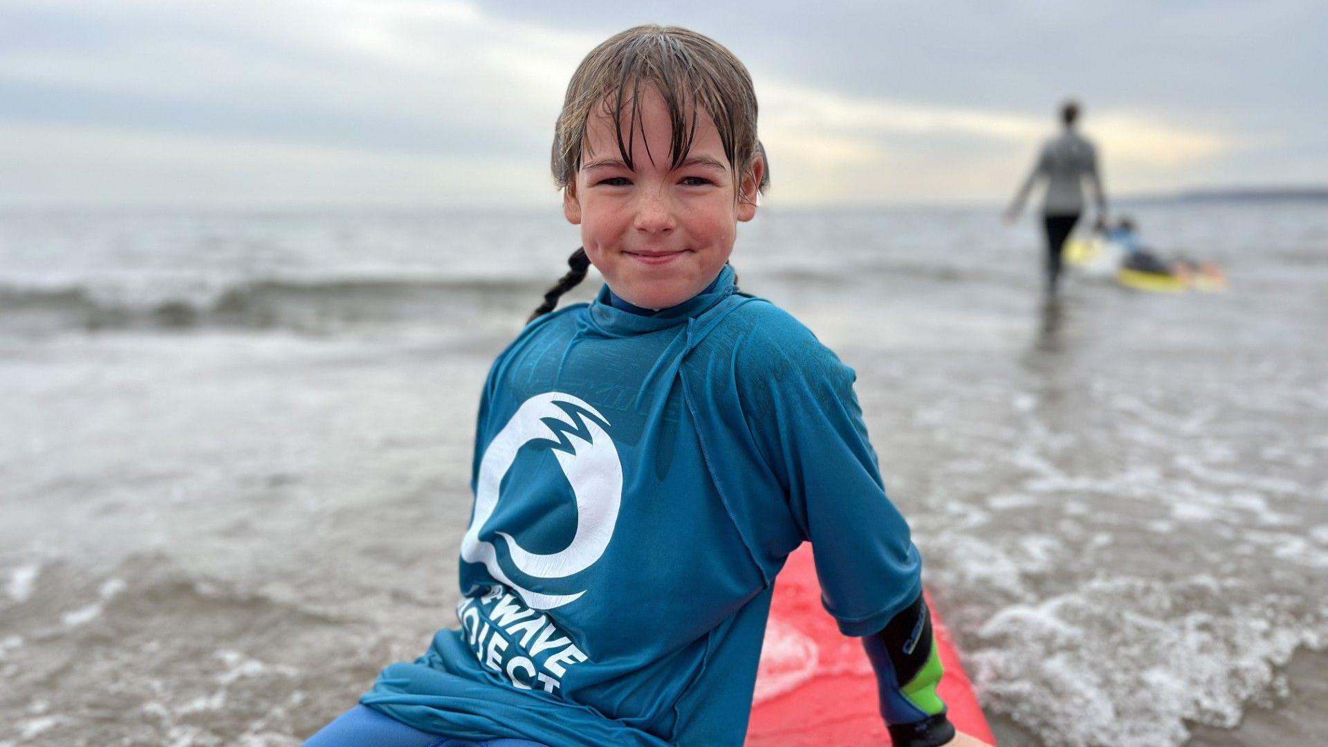 A young girl is sat on a red surfboard in the ocean. It is just where the sand meets the water. Some small waves can be seen. She is wearing a blue top with 'The Wave Project' on it and a wetsuit underneath. She is smiling at the camera. She has brunette hair, a box fringe, and her hair has been put into two plaits.