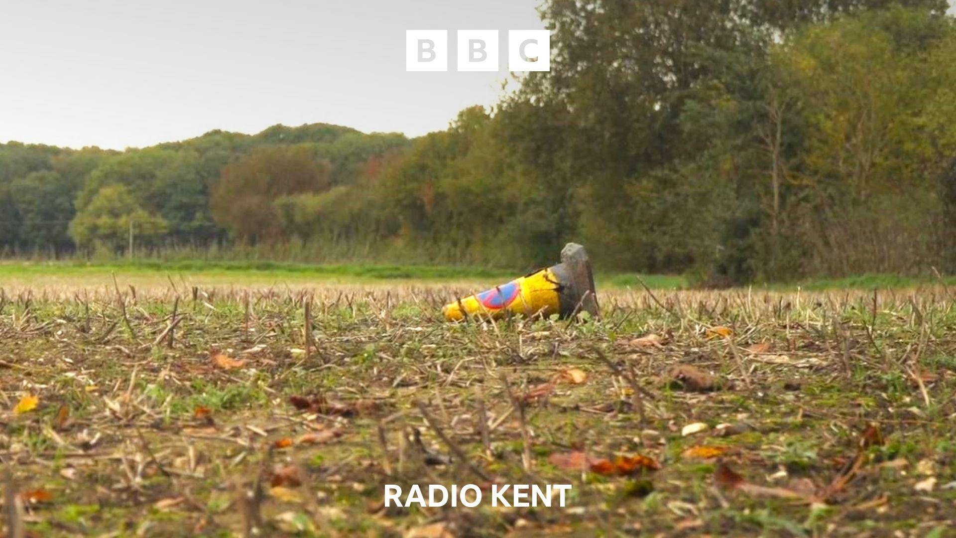 An abandoned traffic cone lies on it's side in the middle of a field.  The field appears as though it was once farmland and is lined with mature trees.