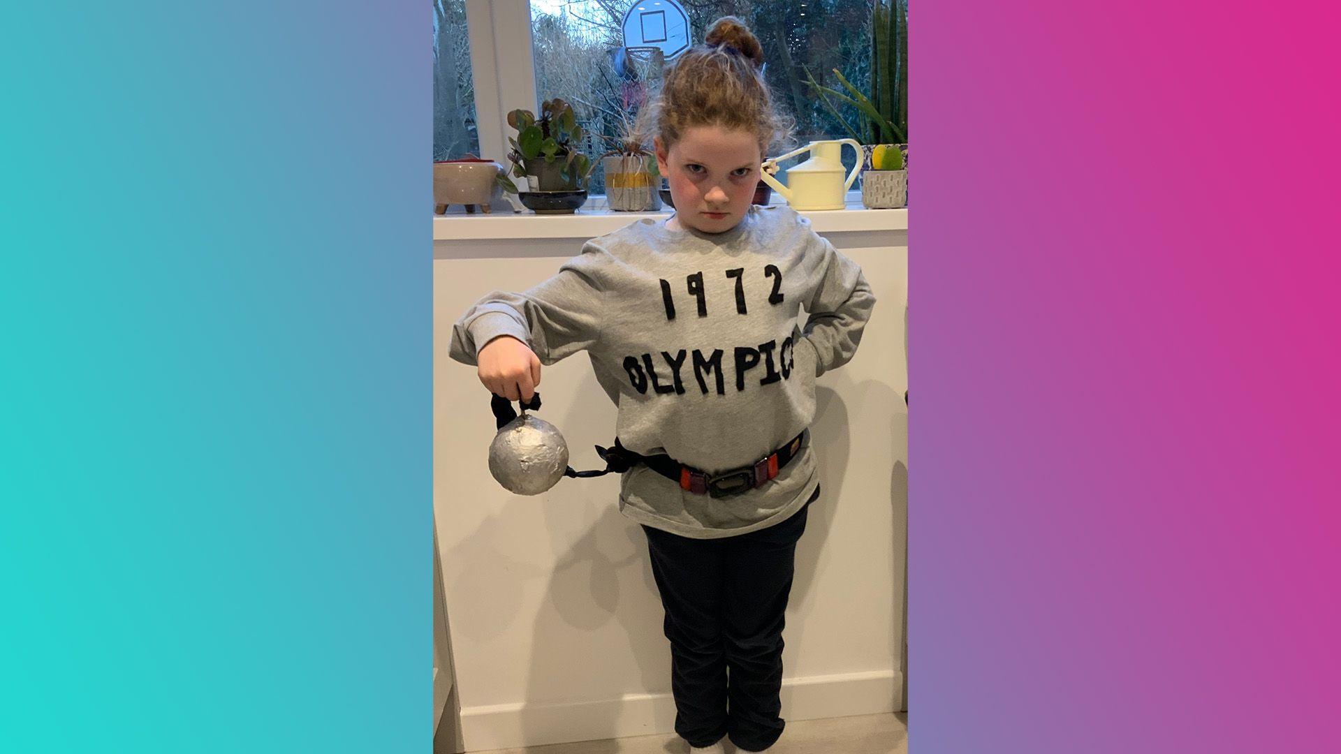 Charlotte has her hair scraped into a bun with a mean stare into the camera. She is wearing black trousers and a grey sweatshirt saying '1972 Olympics' on it and a belt around her middle. She is also carrying a shot put. 